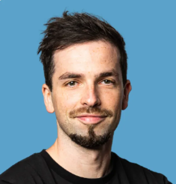 Porträt eines jungen Mannes mit kurzem dunklem Haar, leichtem Bart und schwarzem T-Shirt vor blauem Hintergrund.