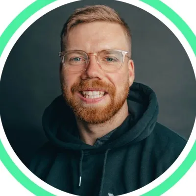 Junger Mann mit kurzem braunem Haar und weißem Pullover lächelt vor weißem Hintergrund.