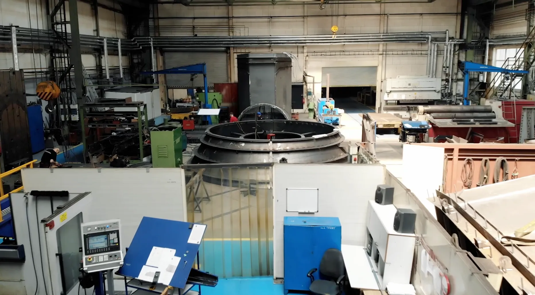 Průmyslový interiér s velkými kovovými zařízeními, modrými skříňkami a pracovní stanicí v popředí.