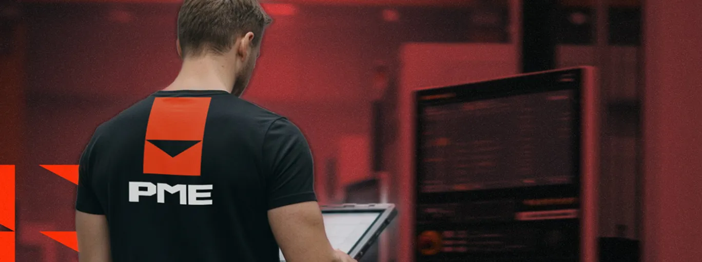 Muž v černém tričku s logem PME čte data na tabletu před obrazovkou s technickými údaji.