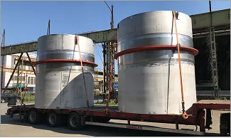 Dvě velké kovové průmyslové nádoby naložené na plošinovém návěsu na otevřeném prostranství.