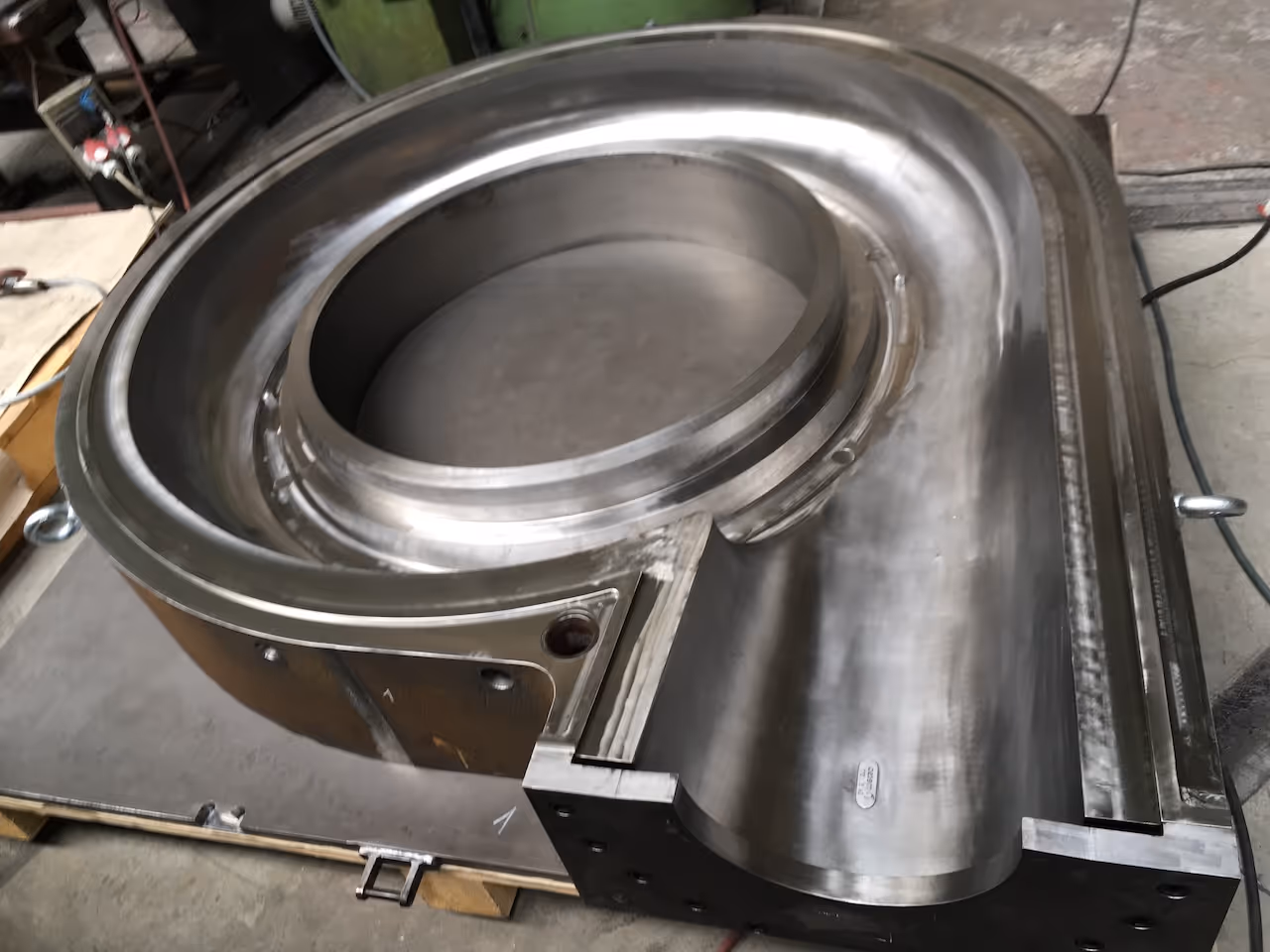 Velký kovový průmyslový součást s kulatým a zakřiveným tvarem na dílním podkladu.