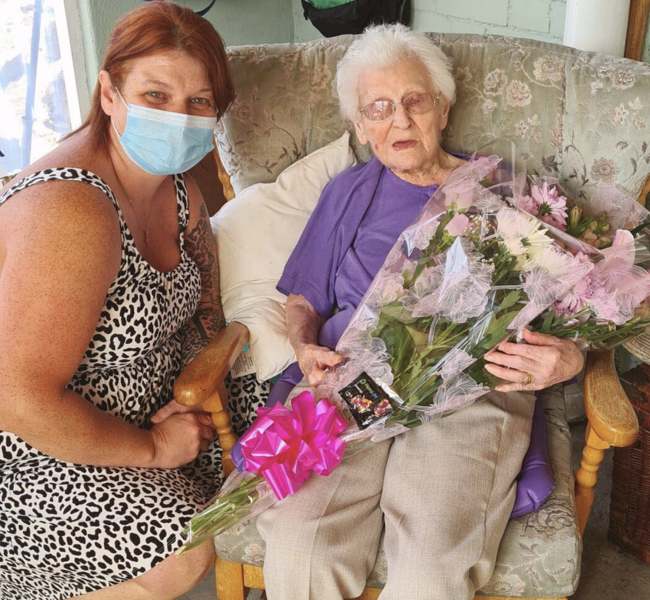 Smiling woman with mask sitting beside elderly woman holding a bouquet wrapped with pink bow.
