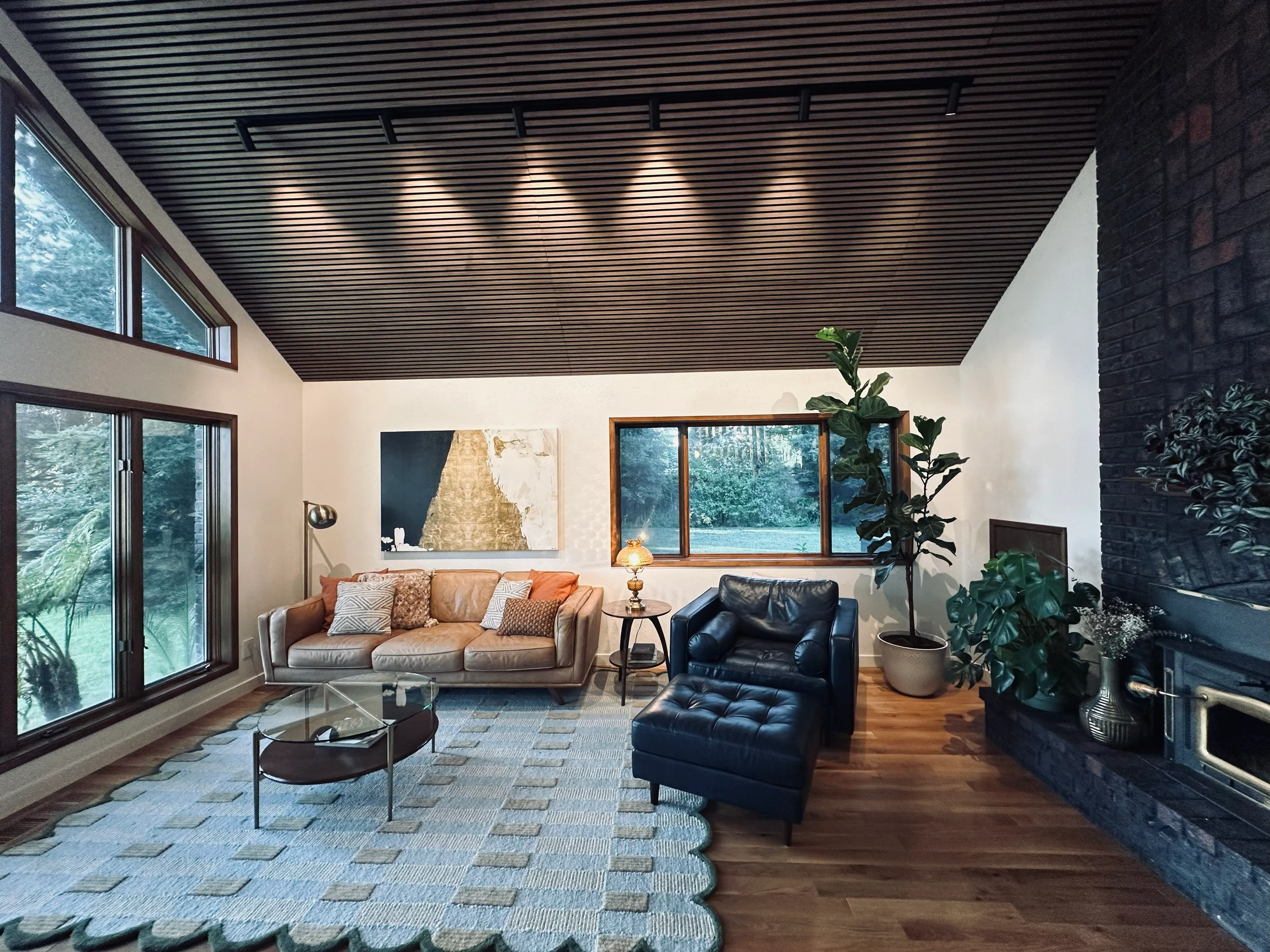 Modern living room with leather sofa, black armchair, large windows, wooden ceiling, and plants near fireplace.