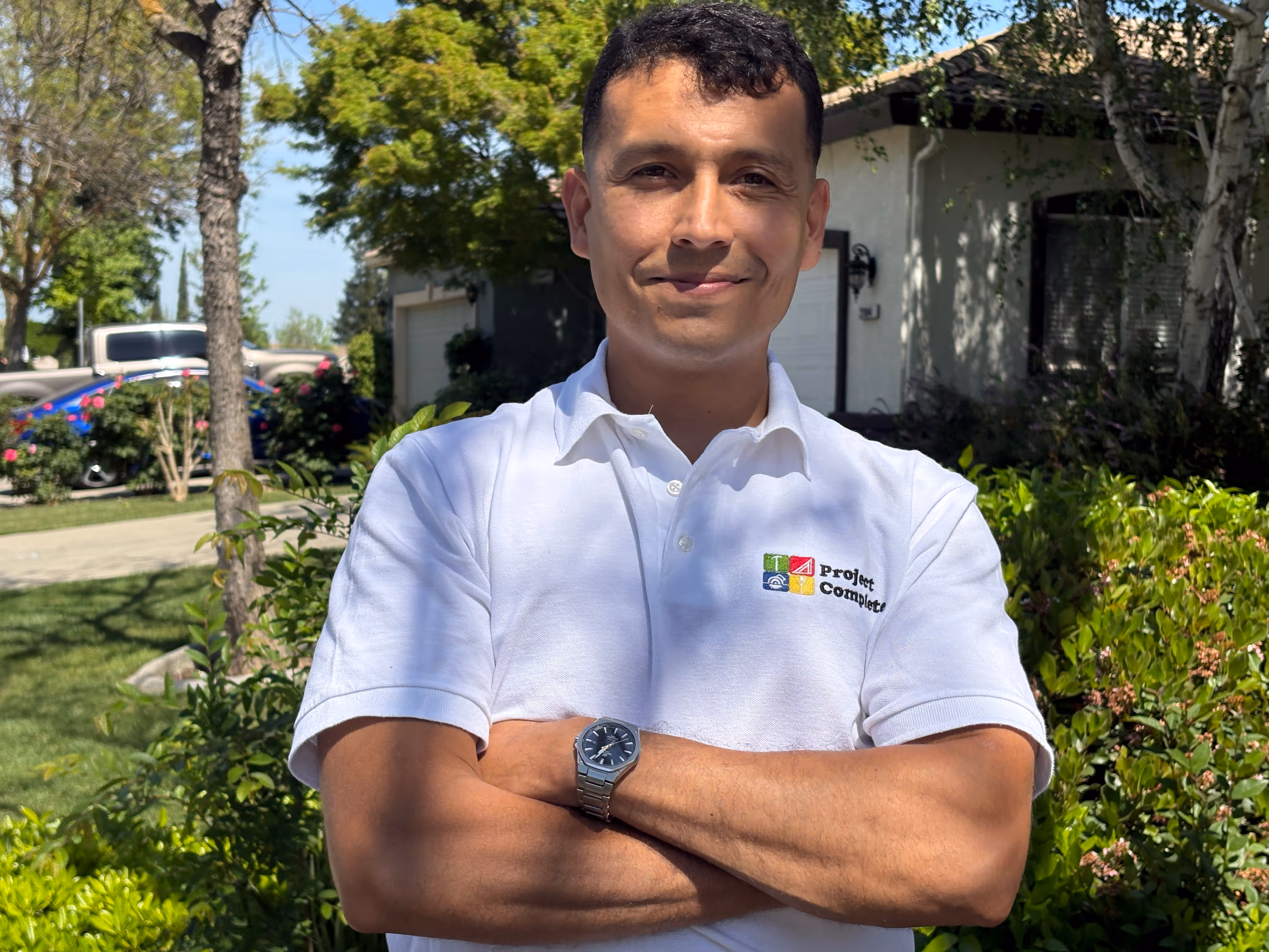 Man standing outdoors with arms crossed wearing a white Project Complete polo shirt.