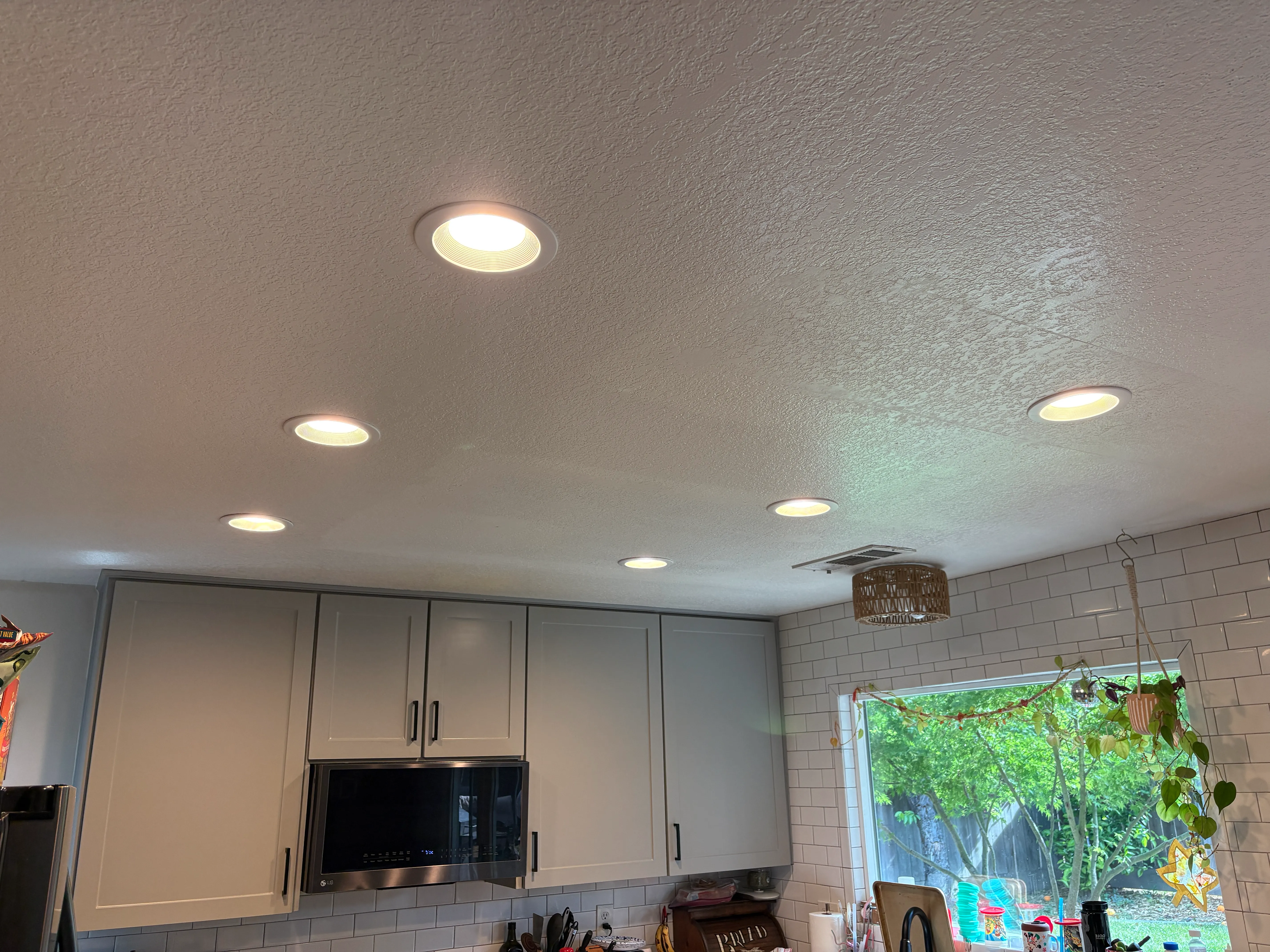 Ceiling with six recessed lights over a kitchen area with cabinets and a window showing greenery outside.
