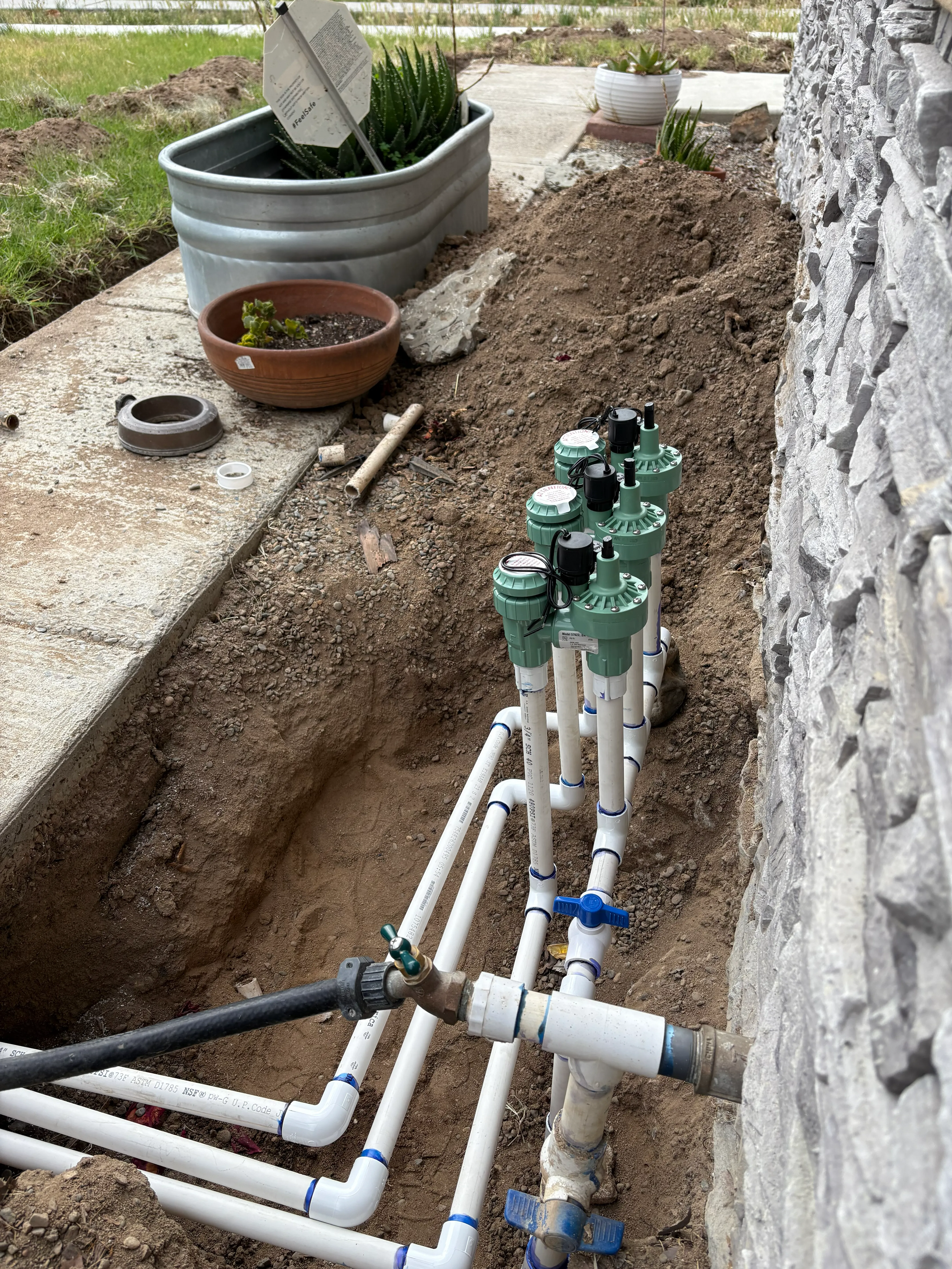 PVC pipes and sprinkler valves installed in a dug trench beside a stone wall near a garden path.
