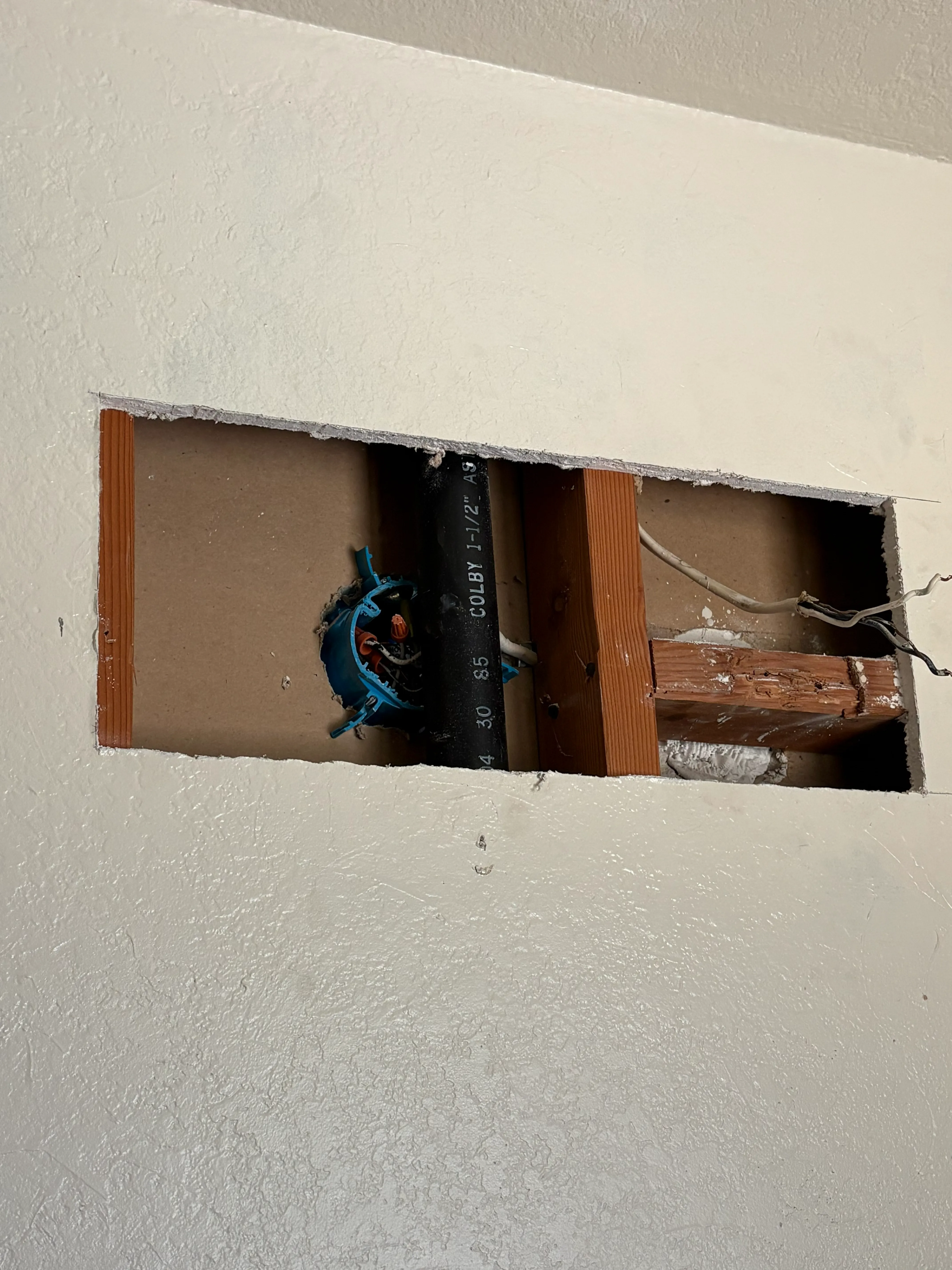 Rectangular drywall cutout revealing wooden studs, electrical box with wires, and a black pipe inside wall.