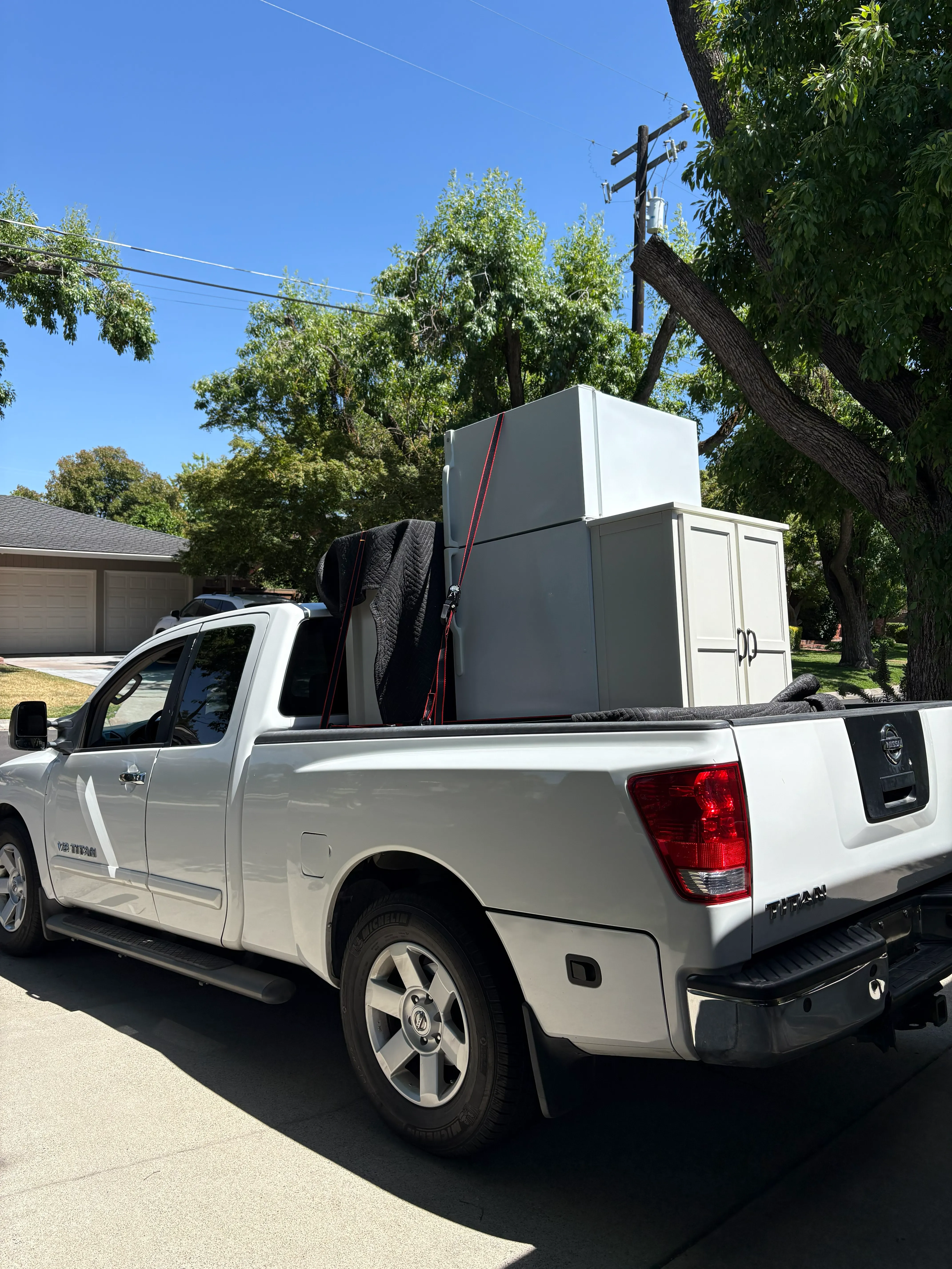 White pickup truck loaded with white appliance boxes and furniture secured with red straps in its bed.