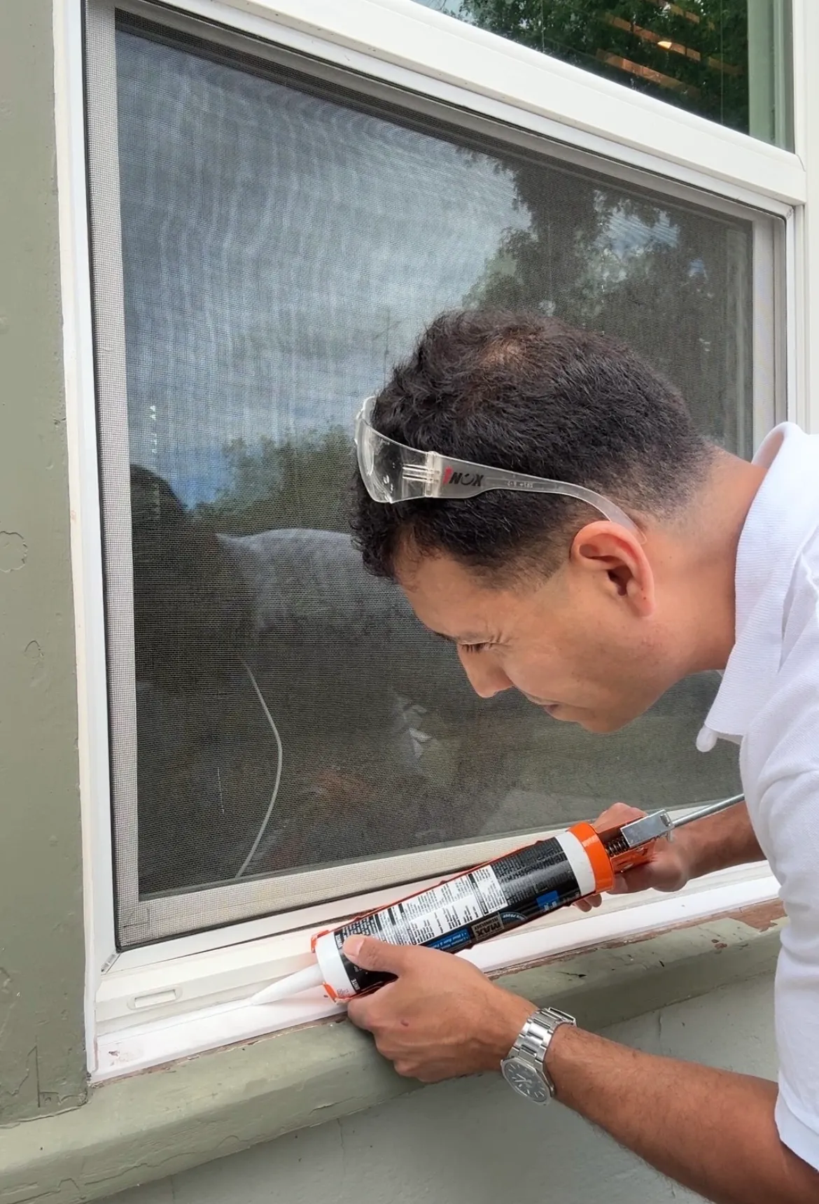 Man wearing safety glasses applying white sealant around a window frame with a caulking gun.