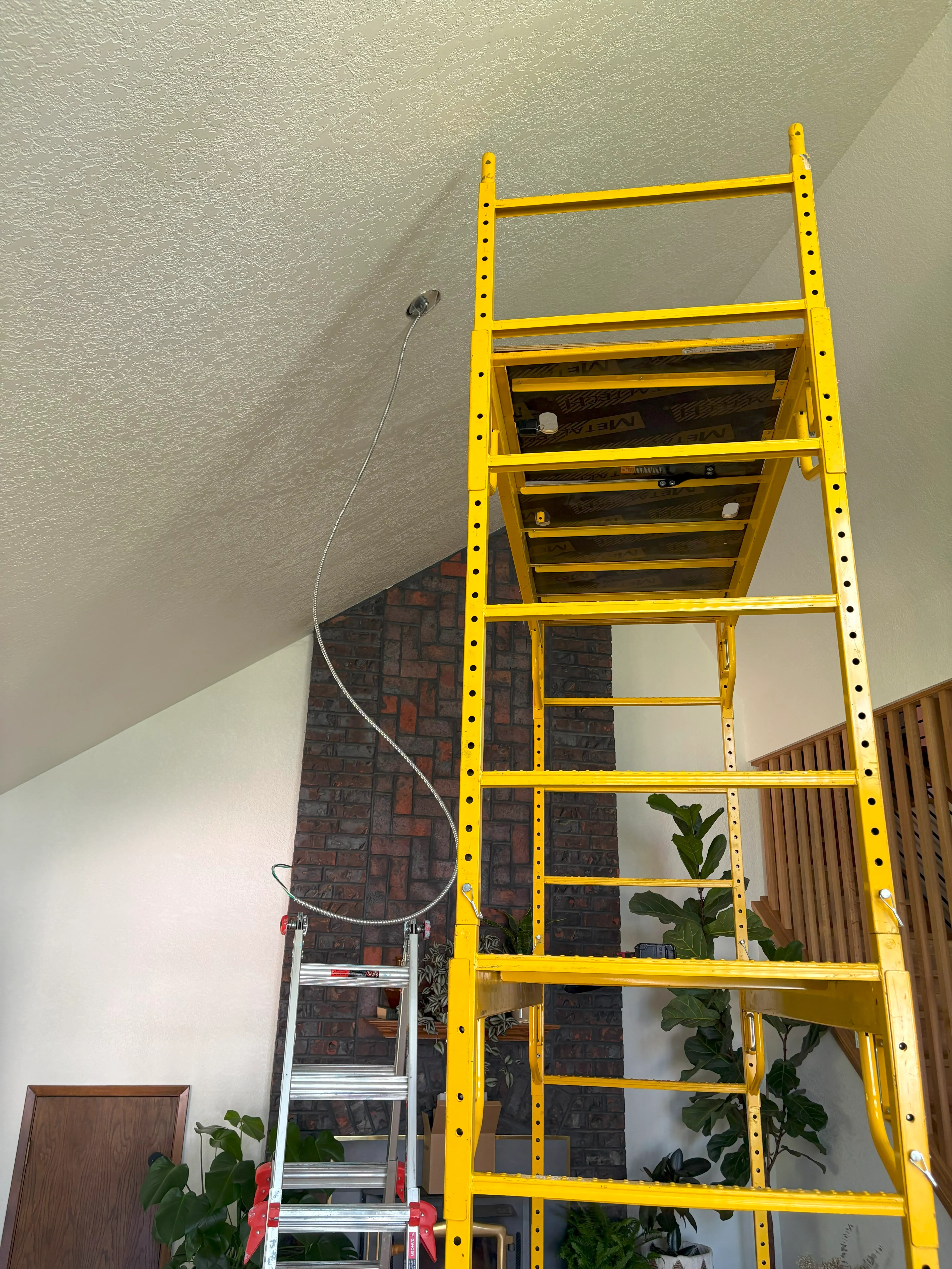 Yellow scaffolding and silver ladder inside room with brick fireplace and plants.