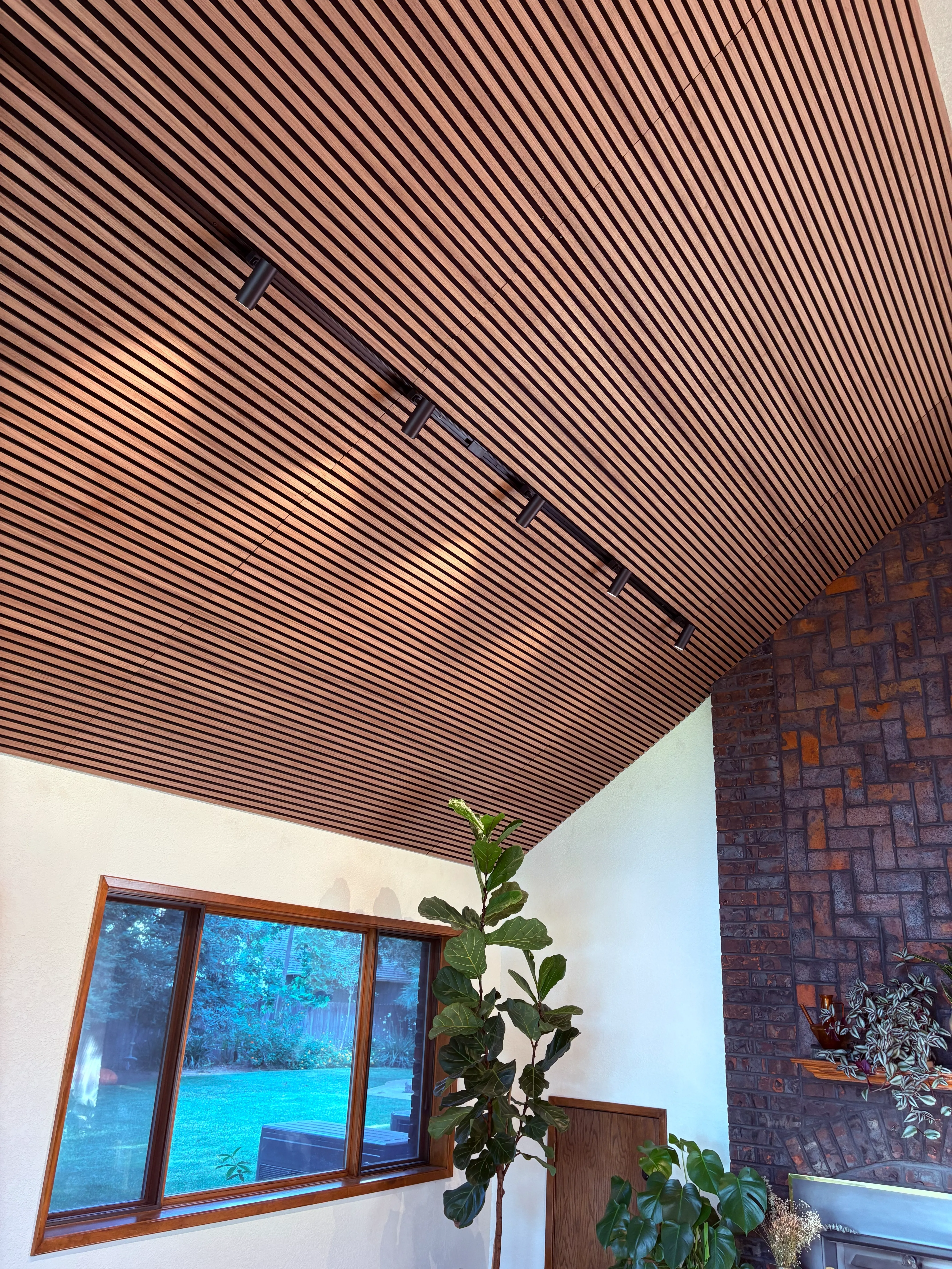 Indoor corner with wood slat ceiling, track lights, large window, brick wall, and tall leafy plants.