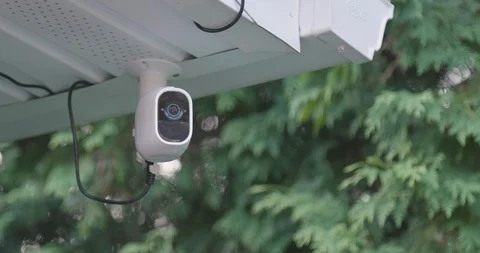 White security camera mounted under roof eave with green trees blurred in background.