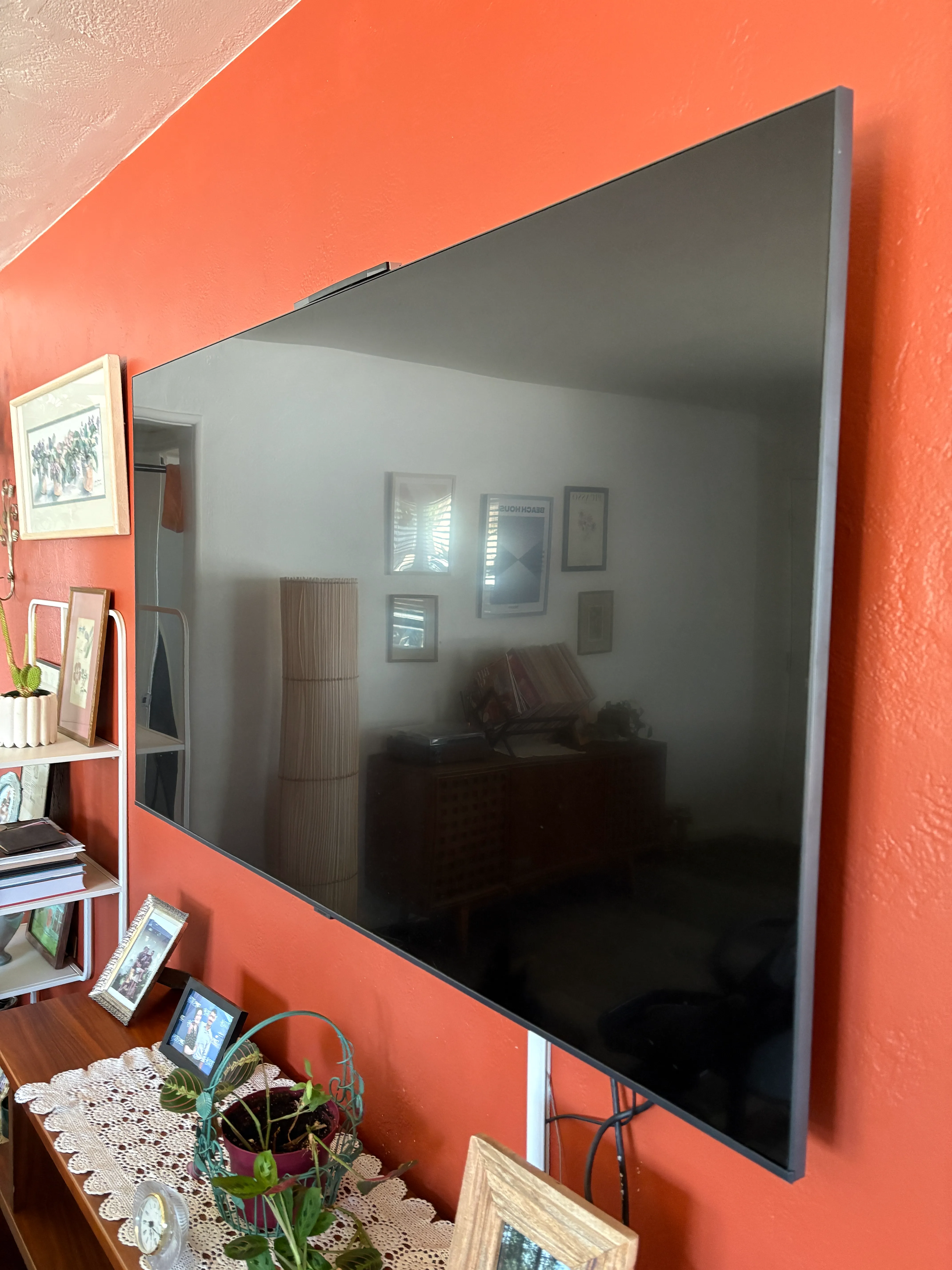 Large flat-screen TV mounted on a textured orange wall above a table with plants and framed photos.