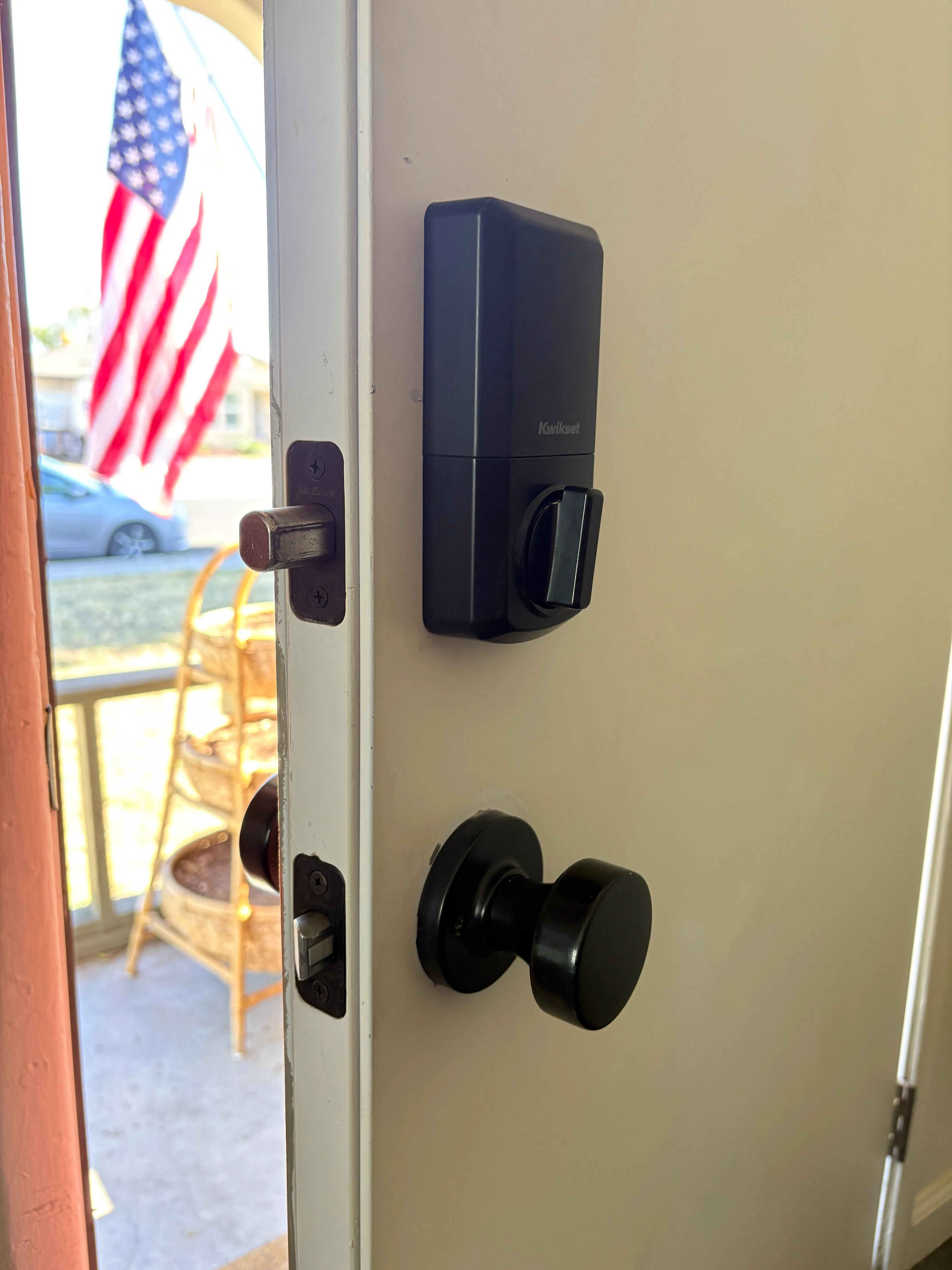 Open white door with black Kwikset smart lock and knob, American flag and chair visible outside.