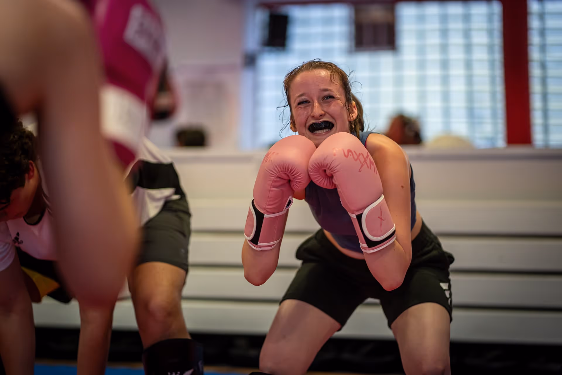 Junge Frau mit rosa Boxhandschuhen und Mundschutz in einem Boxring, die lächelt und sich in Kampfhaltung beugt.