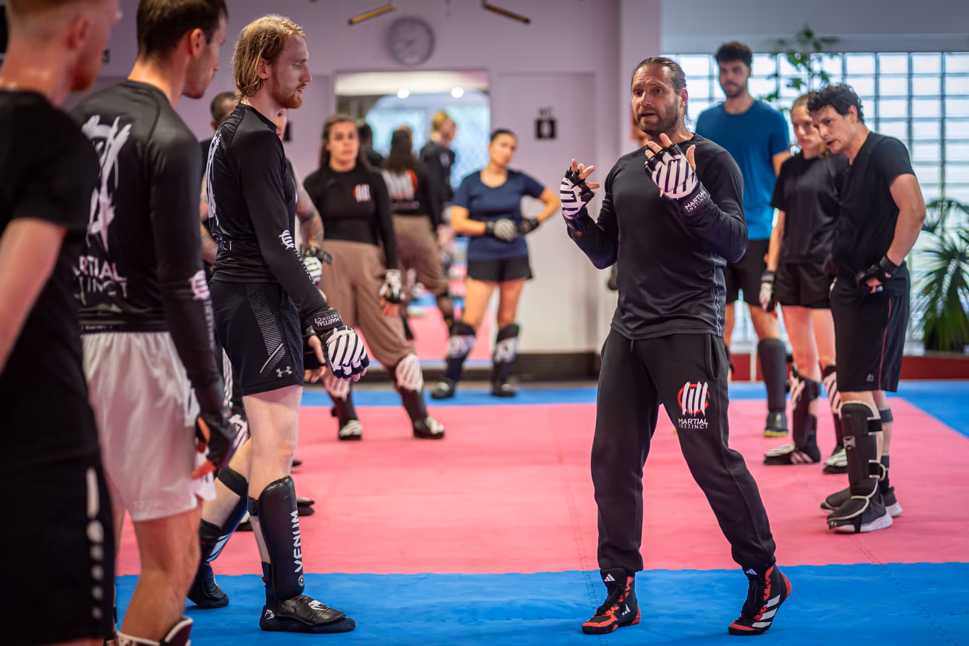 Kampfkunsttrainer erklärt einer Gruppe von Schülern Techniken in einem Trainingsraum mit Mattierung.
