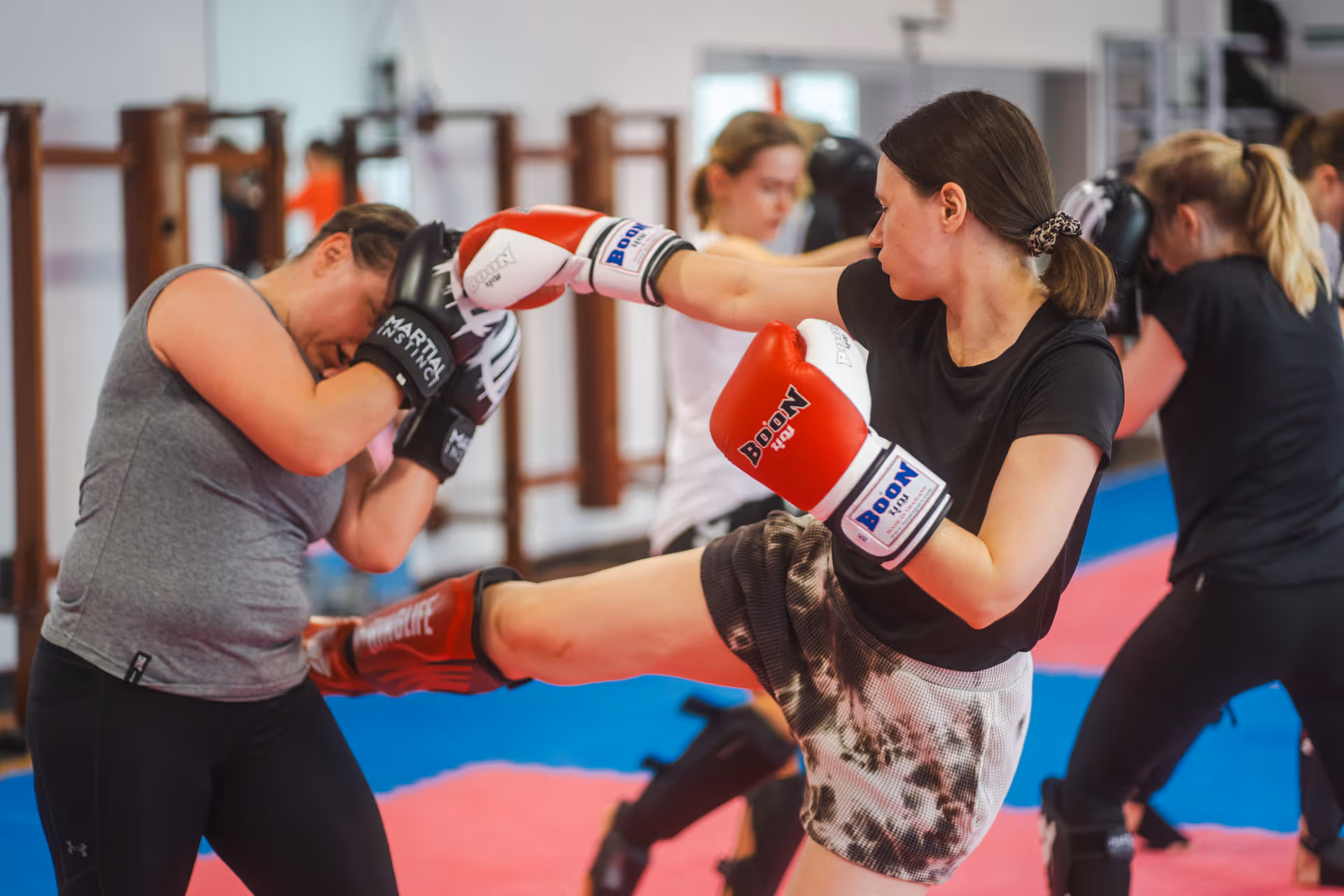 Zwei Frauen trainieren Kampfsport in einem Fitnessstudio, die eine führt einen Tritt aus, die andere hält die Arme schützend vor dem Gesicht.