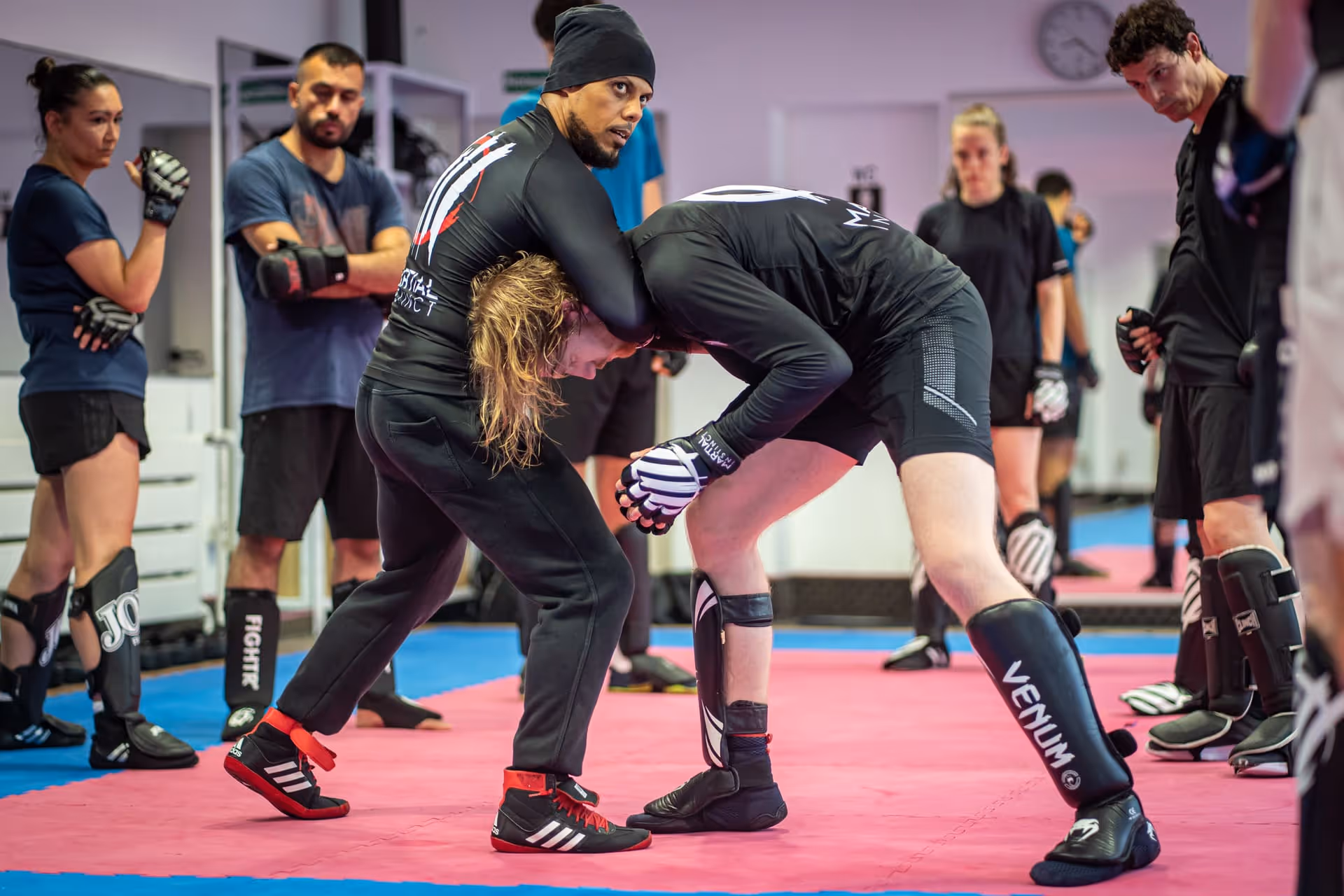 Zwei Männer üben eine Kampfsporttechnik im Training, umgeben von anderen Teilnehmern mit Schutzausrüstung in einem Gym.