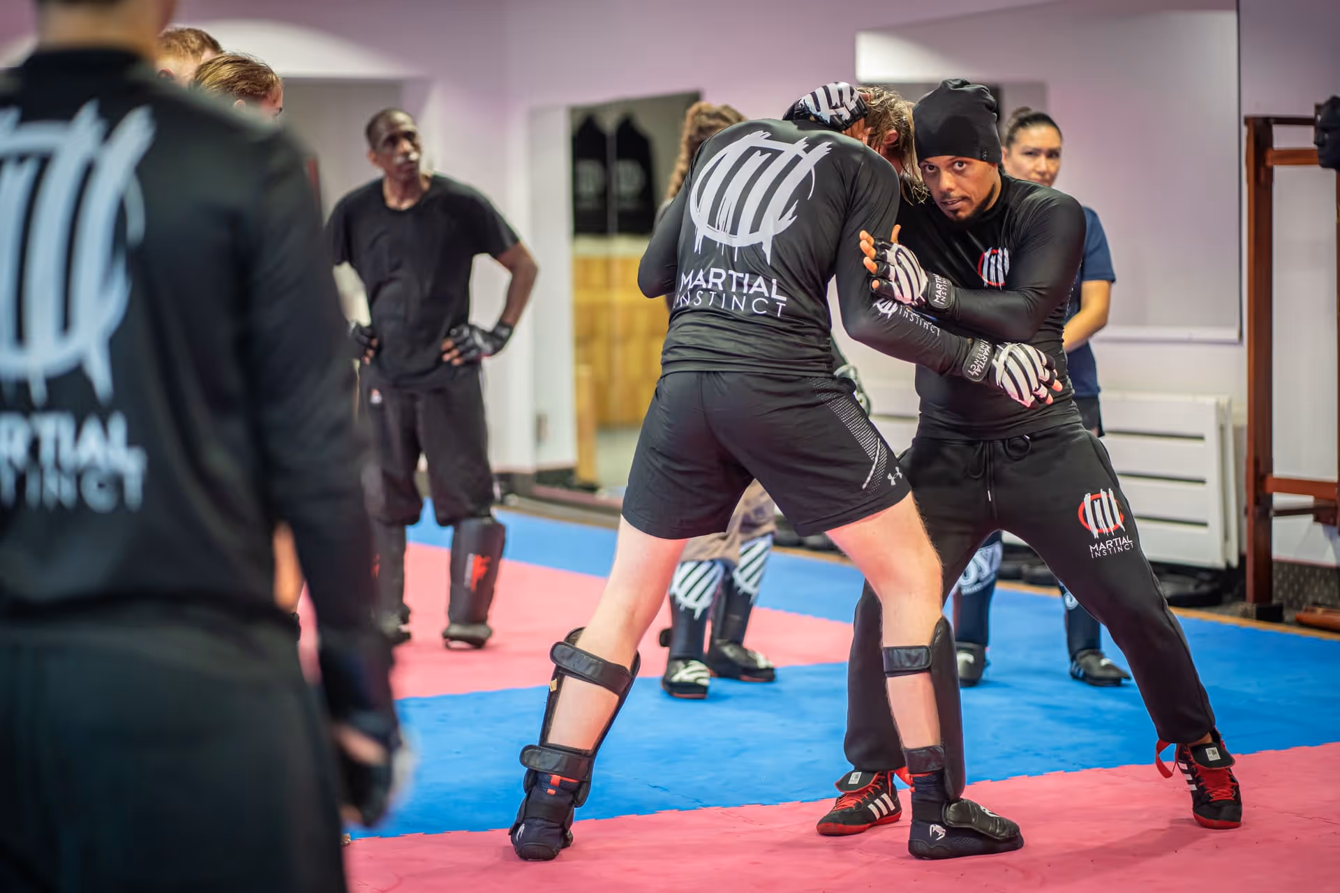 Zwei Männer üben Nahkampftechniken in einem Martial Instinct Kampfsportstudio mit anderen Teilnehmern im Hintergrund.