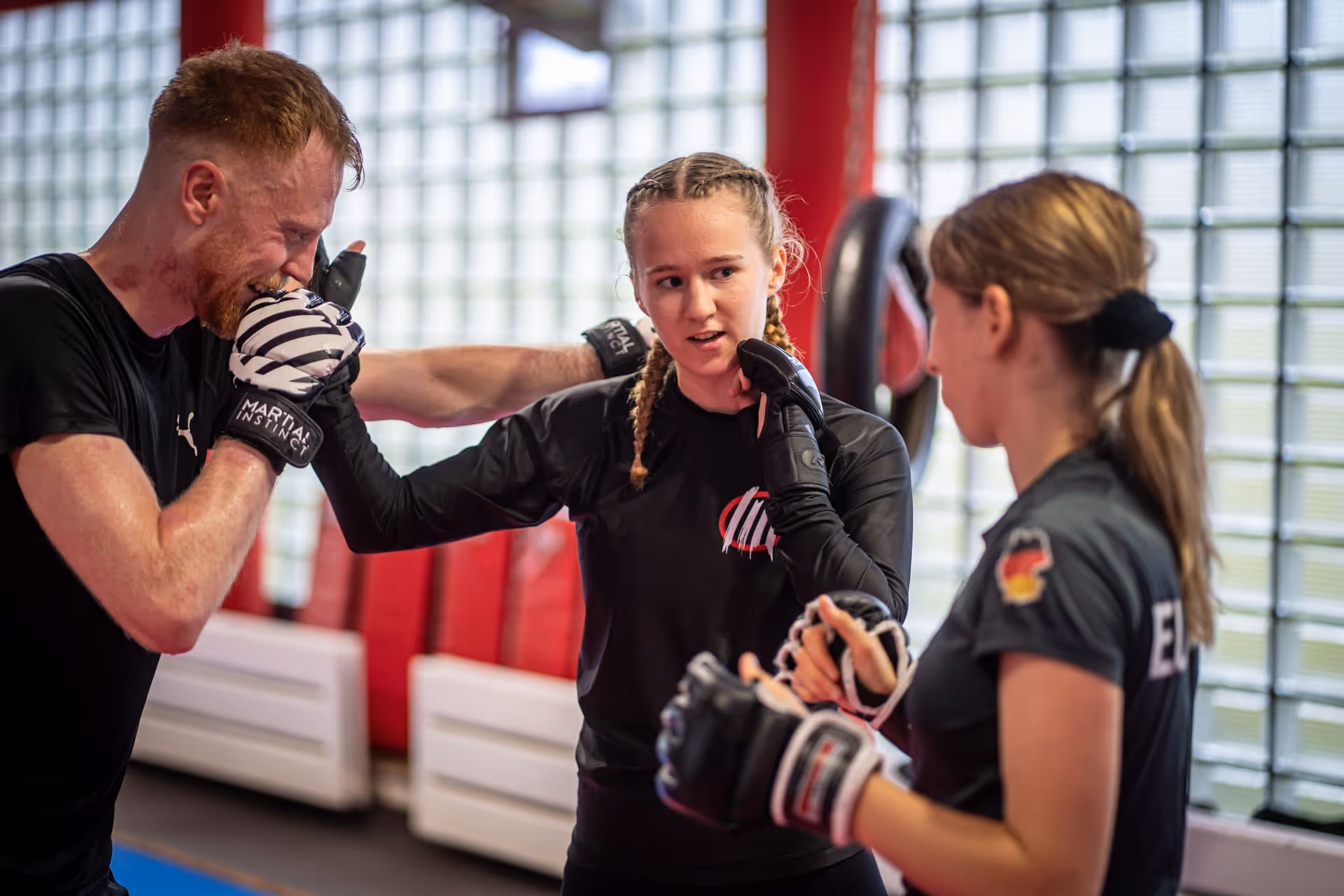 Drei Personen trainieren Mixed Martial Arts in einem hellen Studio, eine Frau zeigt einer anderen Frau und einem Mann eine Technik mit Boxhandschuhen.