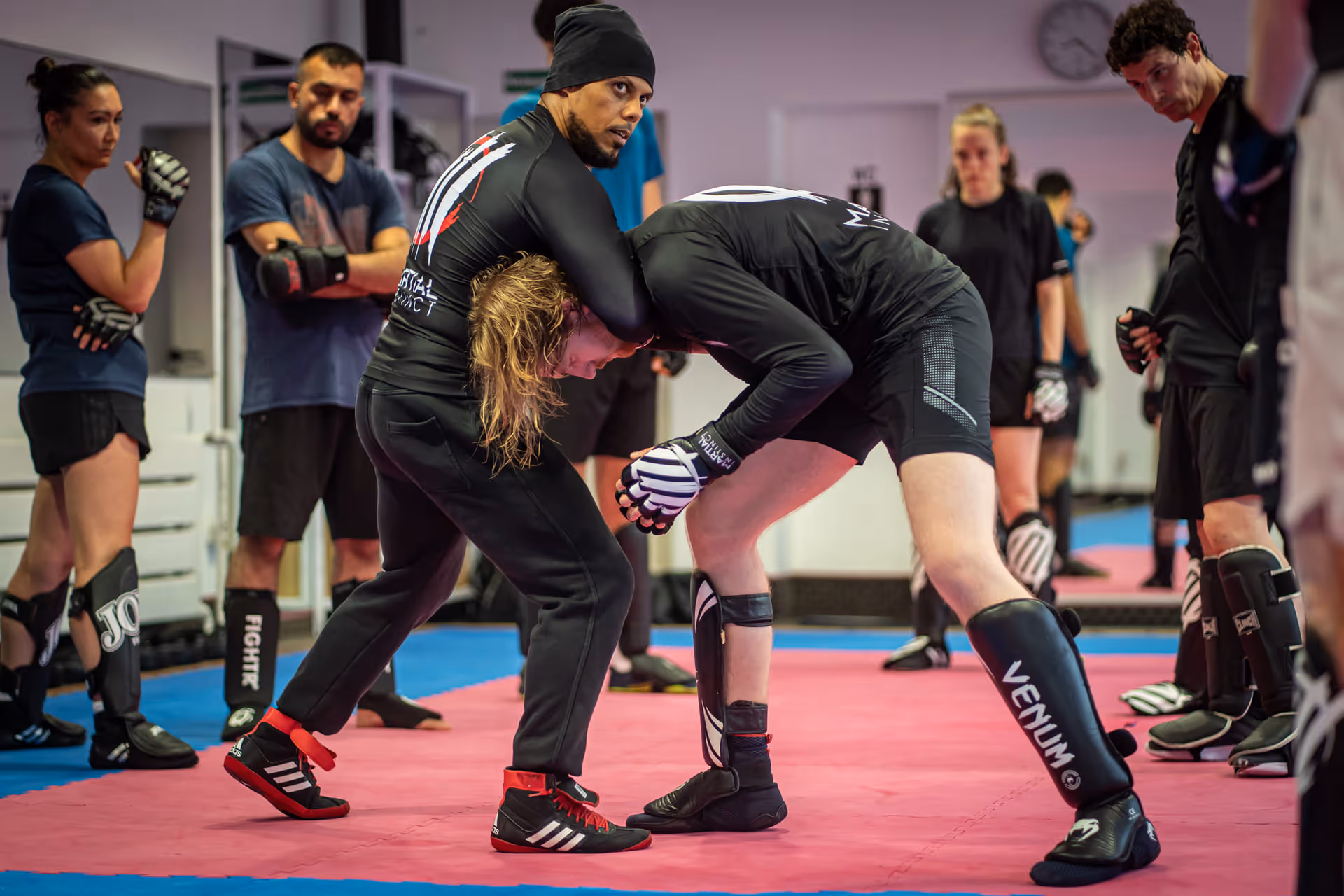 Zwei Männer üben eine Kampfsporttechnik in einem Dojo, während andere Teilnehmer zuschauen.