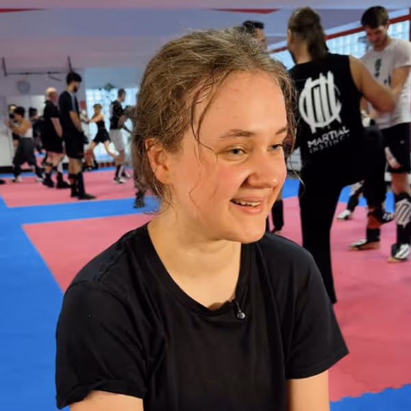 Junge Frau mit zusammengebundenem Haar und schwarzem T-Shirt lächelt in einem Kampfsportstudio mit anderen trainierenden Personen im Hintergrund.