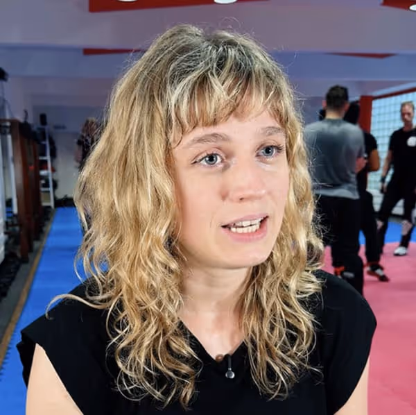 Frau mit lockigen blonden Haaren in schwarzem T-Shirt spricht in einem Gym mit blauem und rotem Boden.