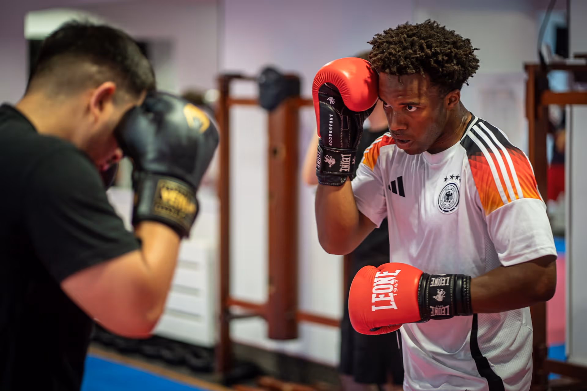 Zwei Männer im Fitnessstudio trainieren Kickboxen, einer trägt rote Boxhandschuhe und ein weißes Trikot mit deutschen Farben.