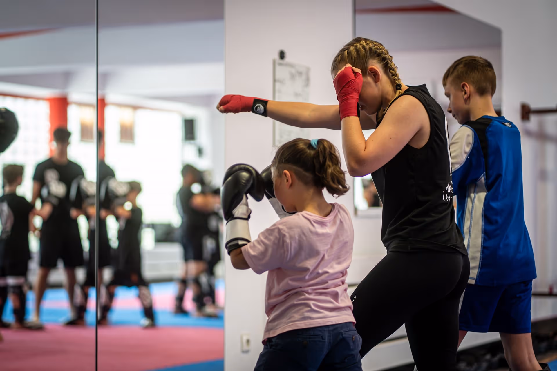 Trainerin mit roten Bandagen trainiert mit einem Mädchen in Boxhandschuhen in einem Fitnessstudio, im Hintergrund spiegelt sich eine Trainingsgruppe.