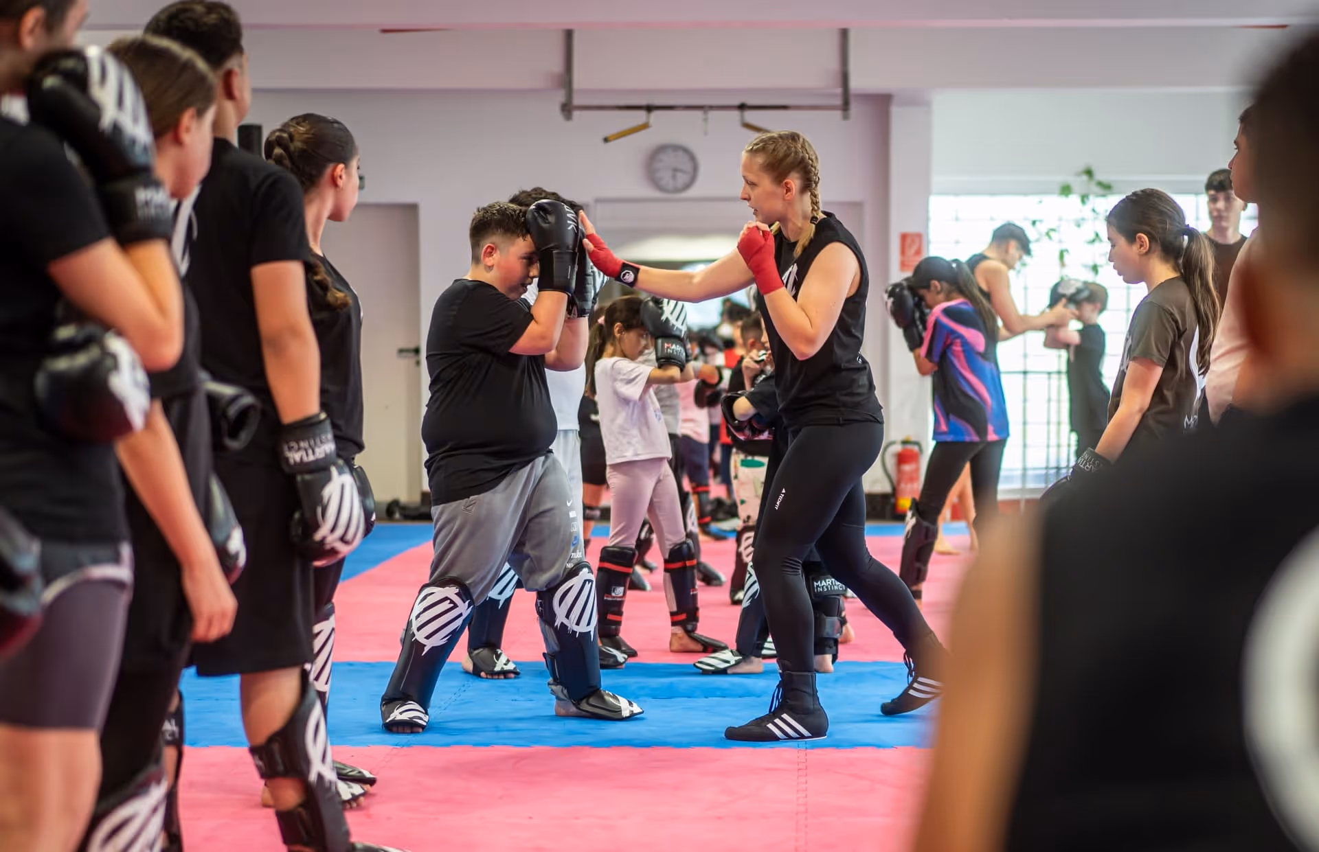 Kinder und Jugendliche trainieren Kickboxen in einer Halle, eine Trainerin gibt Anweisungen.