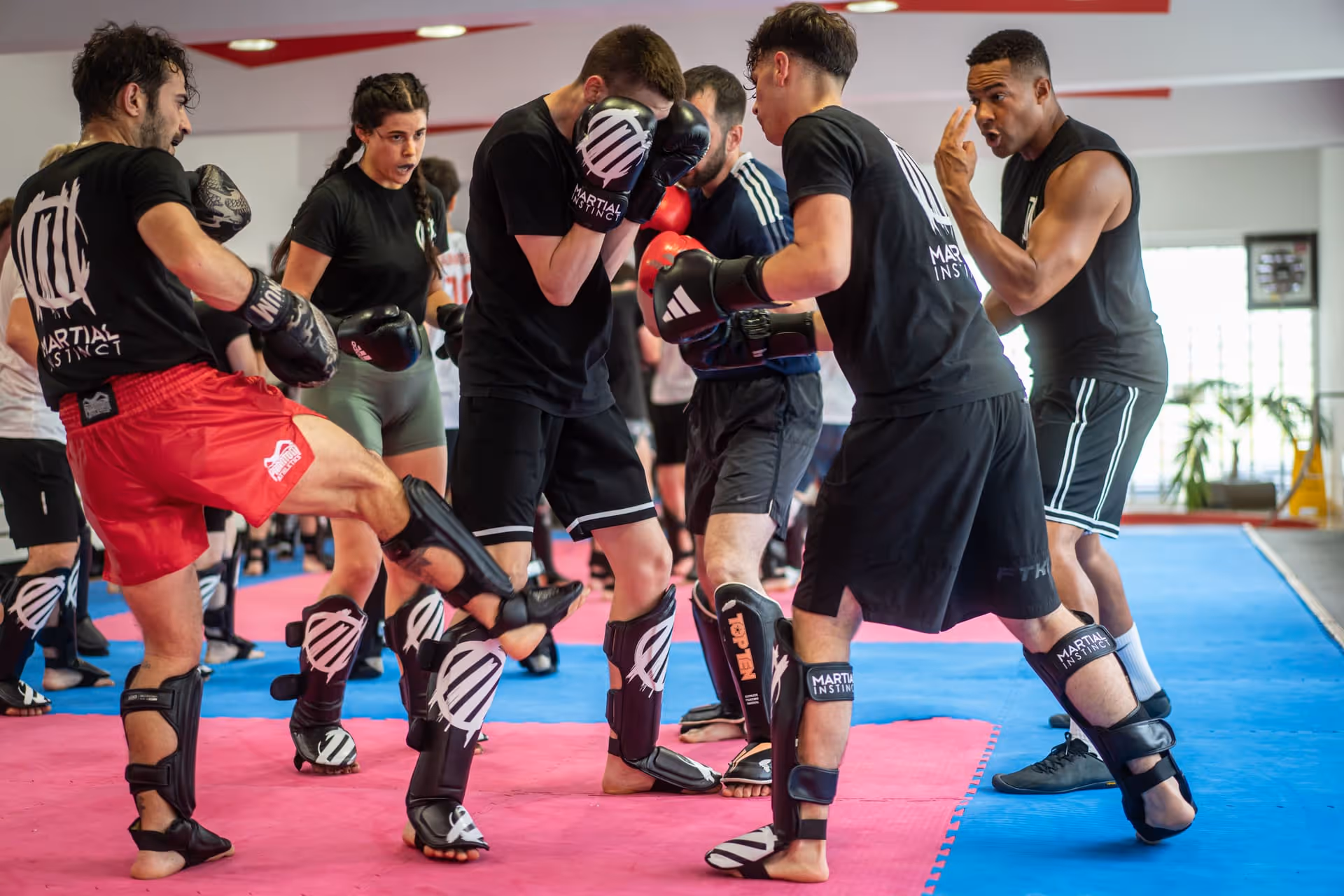 Sechs Personen trainieren zusammen Kickboxen in einem Fitnessstudio mit roten und blauen Matten.