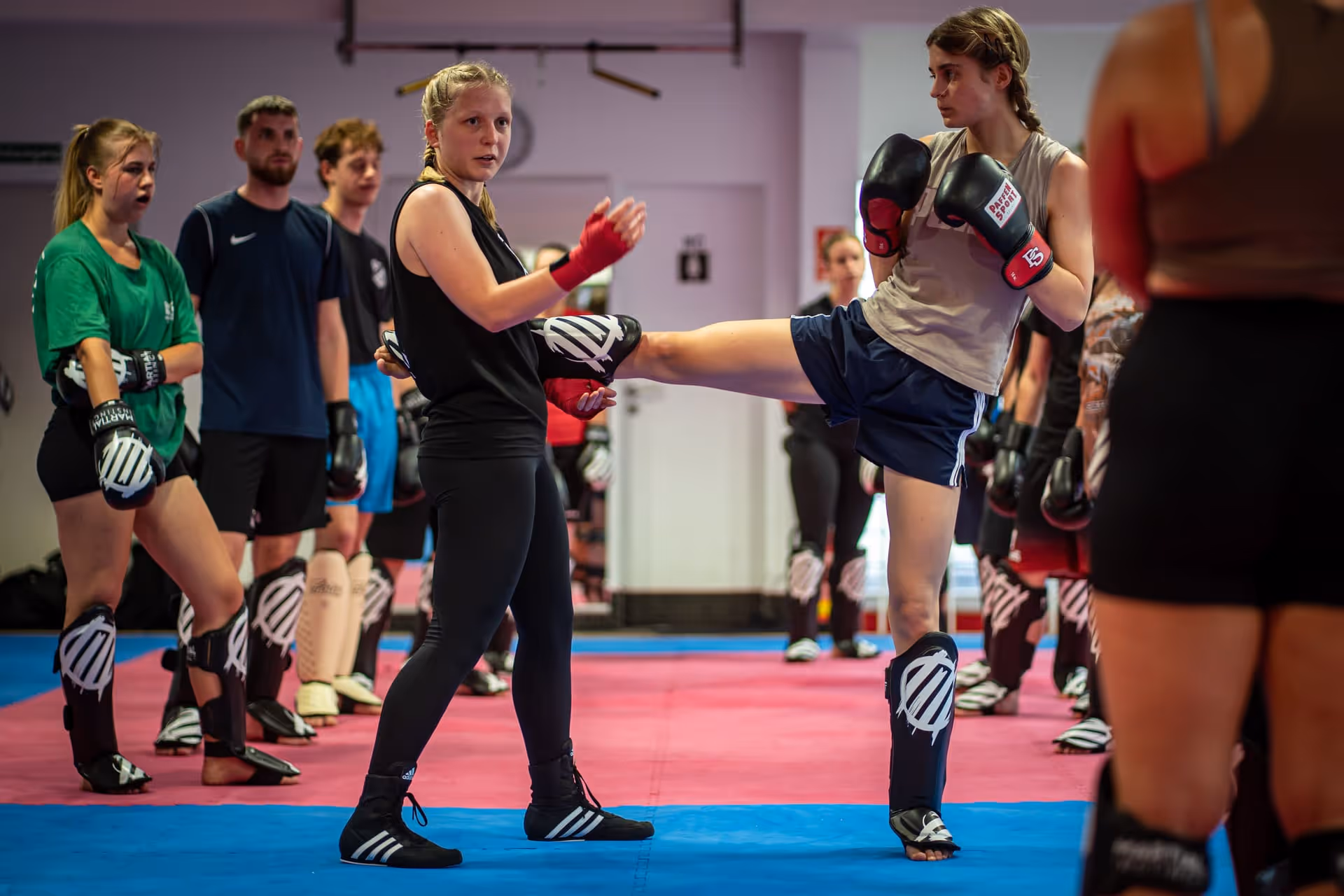 Frauen üben Kickboxen in einer Trainingshalle, eine Frau hält das Bein der trainierenden Frau mit Boxhandschuhen und Schienbeinschonern.