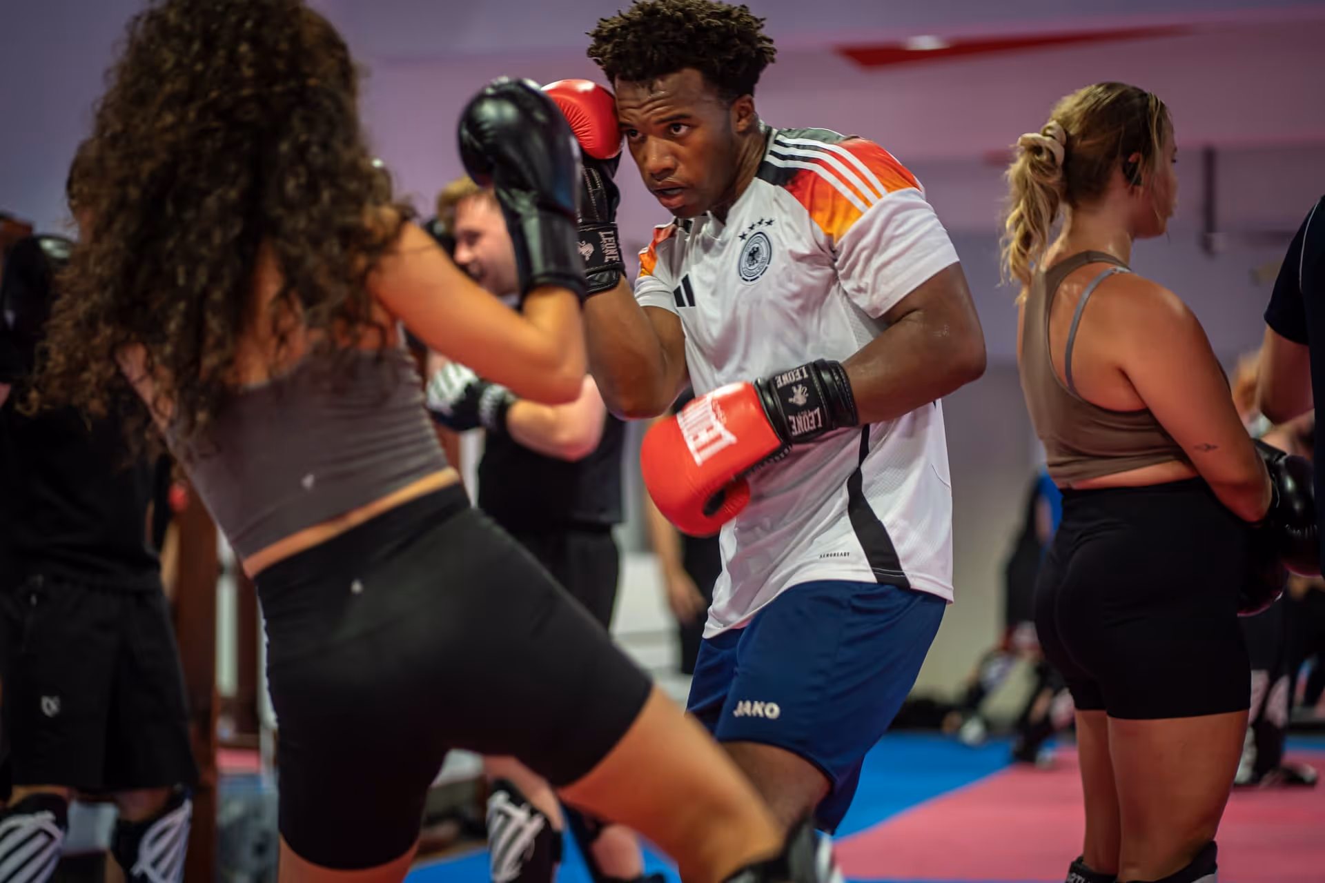 Zwei Menschen trainieren Kickboxen in einem Fitnessstudio, der Mann trägt rote Boxhandschuhe und ein weißes Trikot.