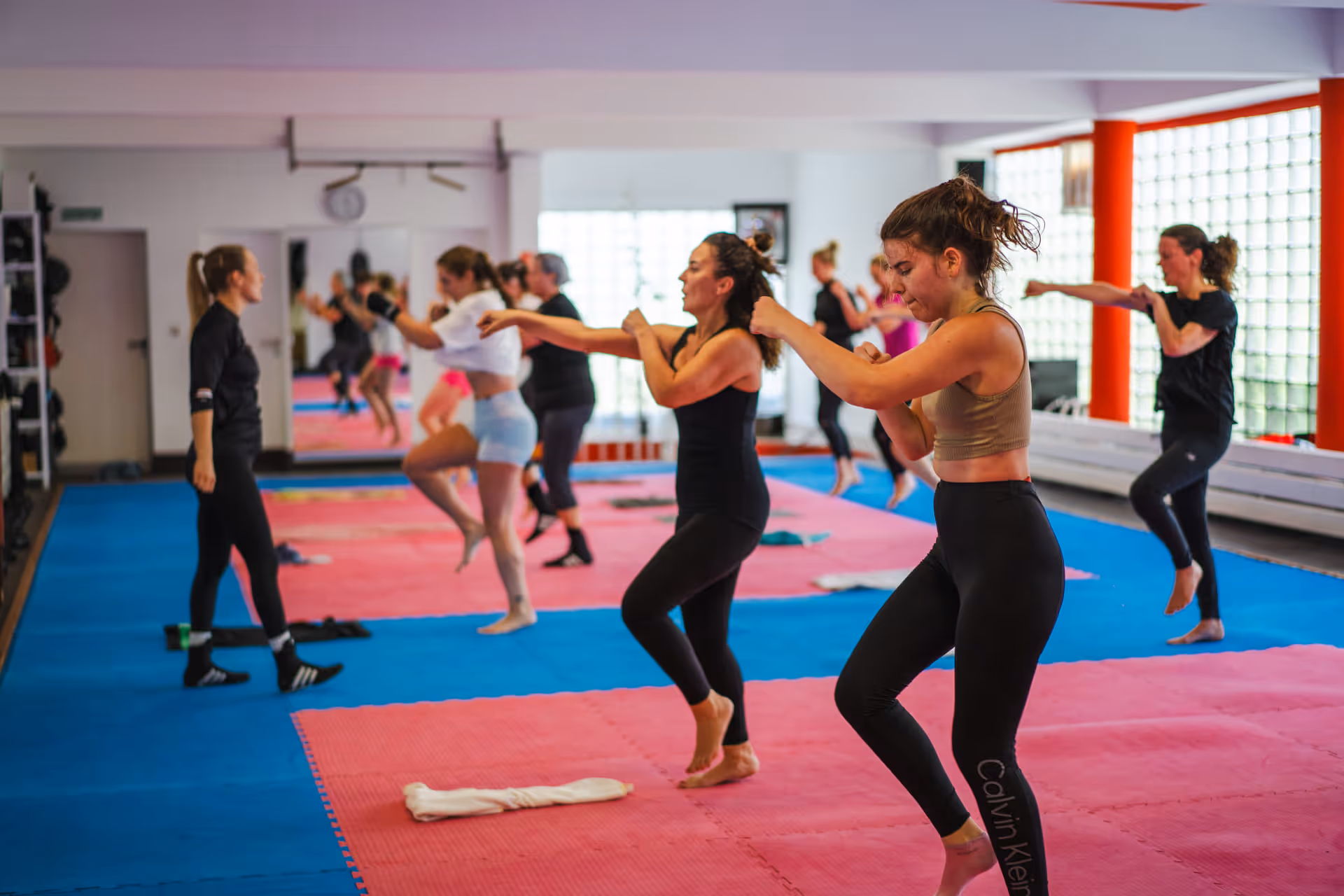 Gruppe von Frauen bei einem Kickbox-Training in einem hellen Fitnessstudio auf blauen und rosa Matten.