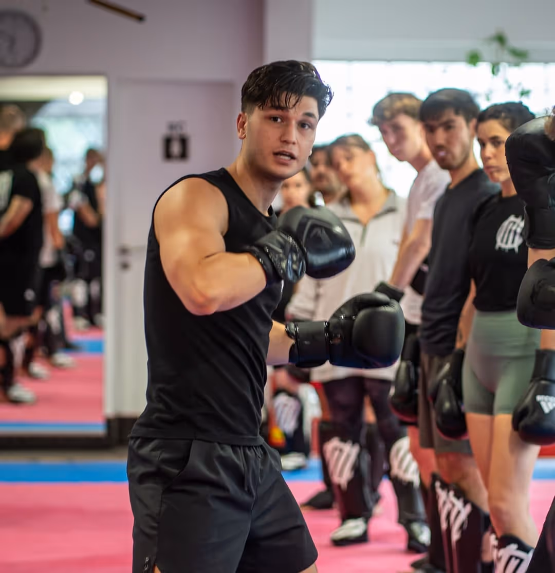 Ein junger Mann in schwarzer Sportkleidung mit Boxhandschuhen zeigt eine Boxhaltung im Vordergrund, während eine Gruppe von Menschen im Hintergrund eine Linie bildet und ebenfalls Boxhandschuhe trägt.