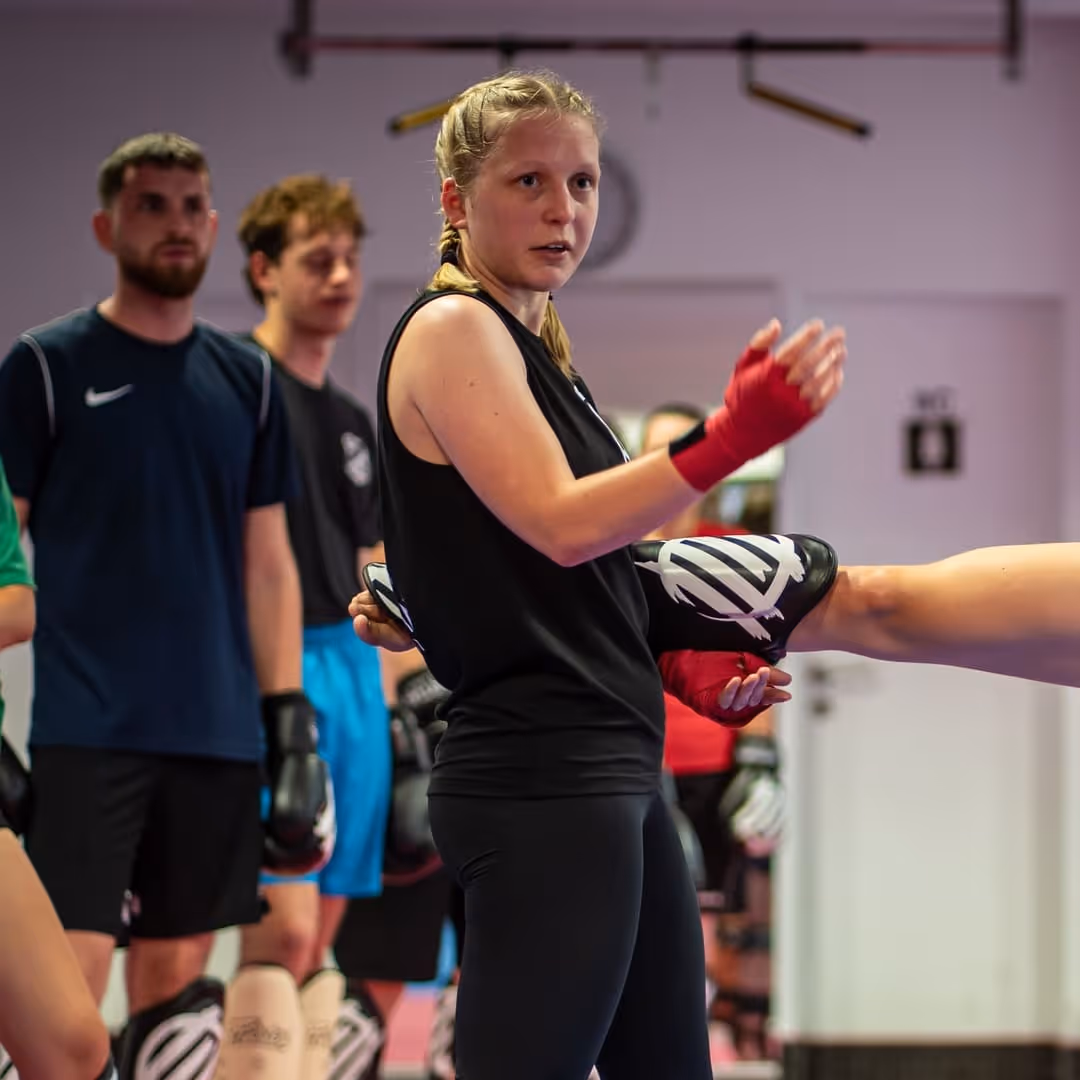 Frau mit roten Handschützern hält Kickpolster, während Person einen Kick in einem Trainingsraum ausführt, mehrere Personen im Hintergrund.