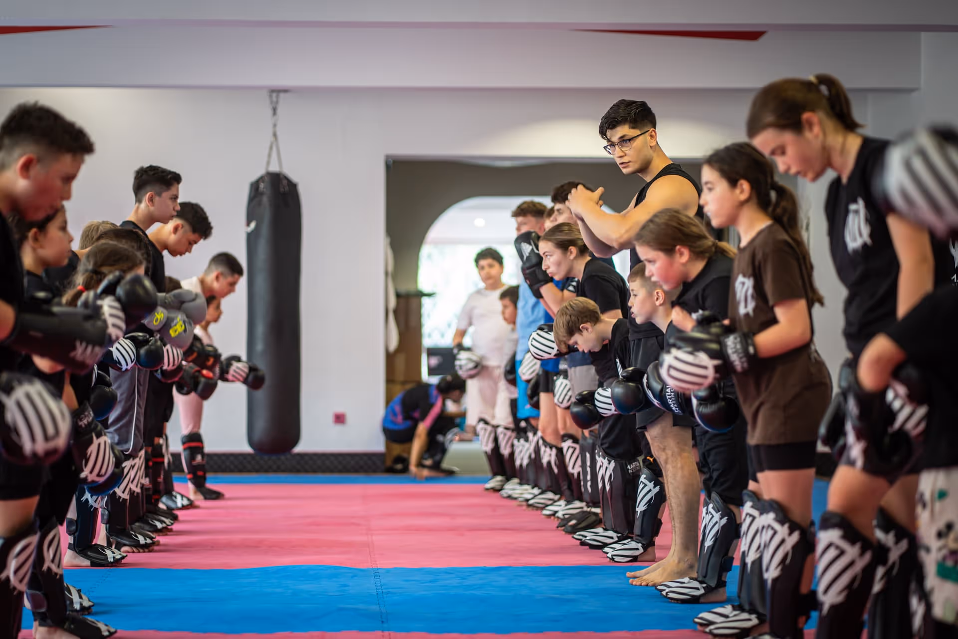 Jugendliche Kampfsportler mit Schutzausrüstung in Kampfstellung auf blauen und pinken Matten in einem Trainingsraum.