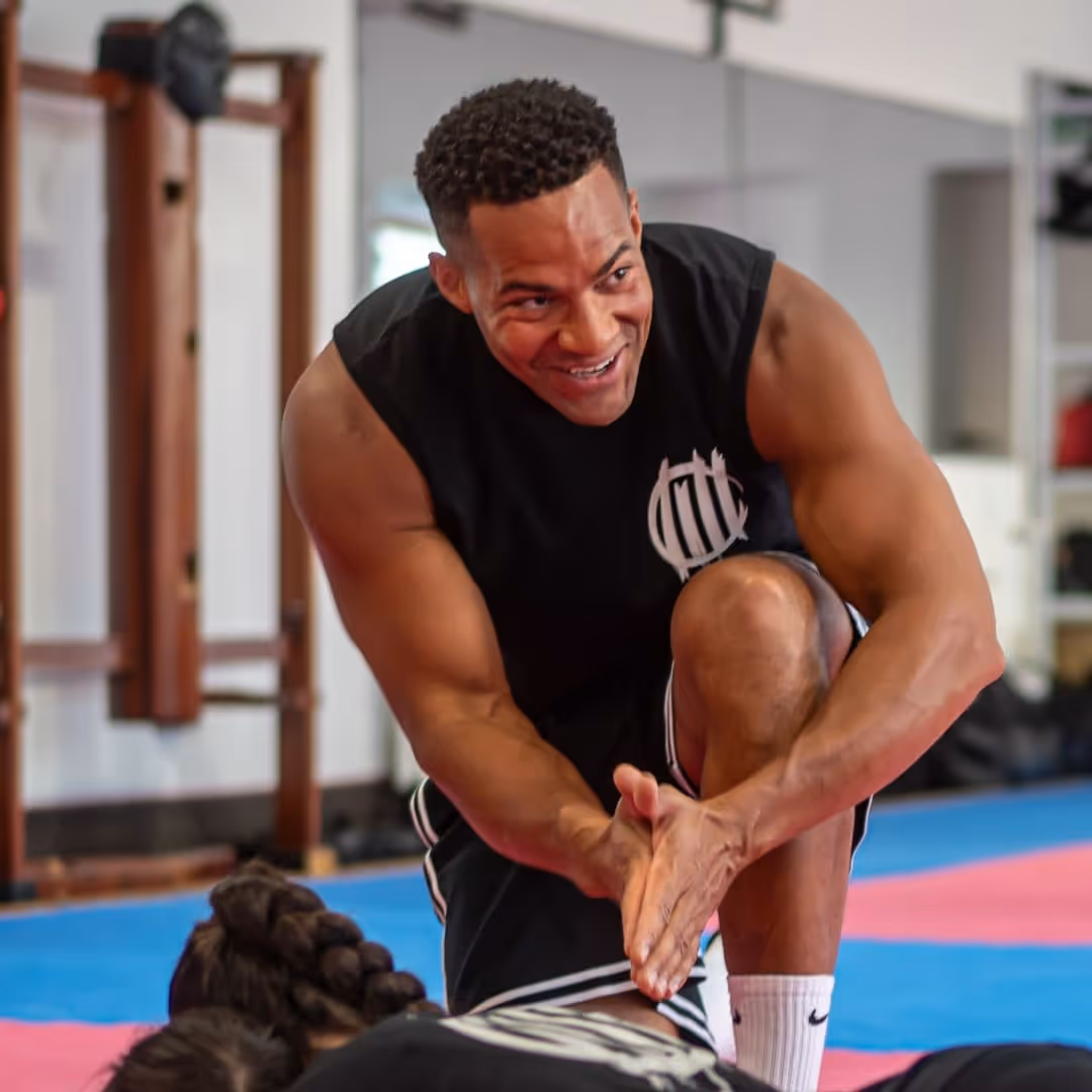 Muskulöser Mann in schwarzem ärmellosen Shirt klatscht in die Hände vor einer Person auf dem Boden in einem Trainingsraum.