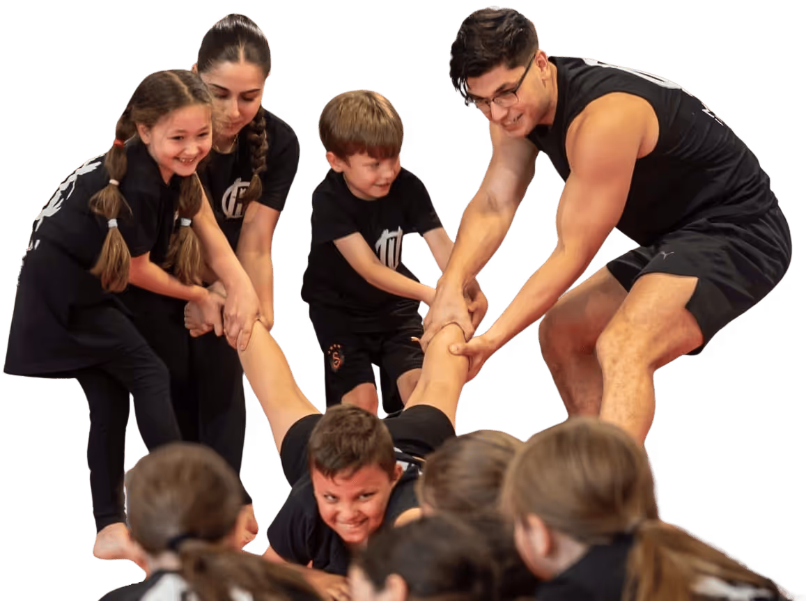 Eine Gruppe von Kindern und einem Erwachsenen hält gemeinsam die Beine eines lächelnden Kindes, das auf dem Boden liegt.