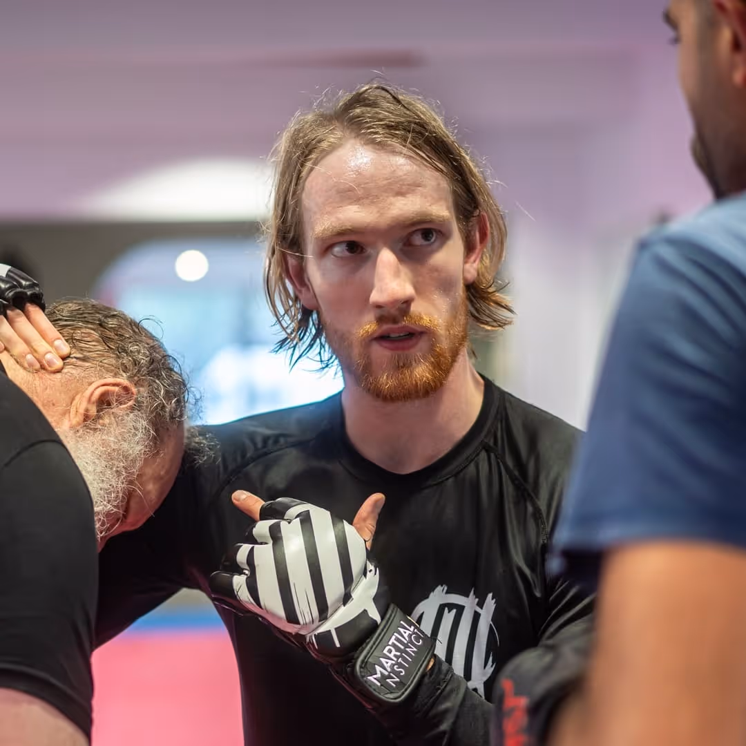 Ein junger Mann mit rotem Bart trägt Handschuhe, hält einen älteren Mann während eines Kampfsporttrainings.