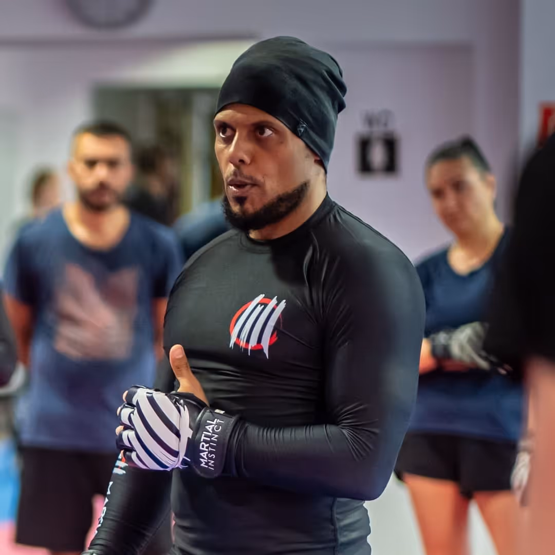 Mann mit schwarzer Sportkleidung und Handschuhen spricht in einem Martial-Arts-Training, mit anderen Teilnehmern im Hintergrund.