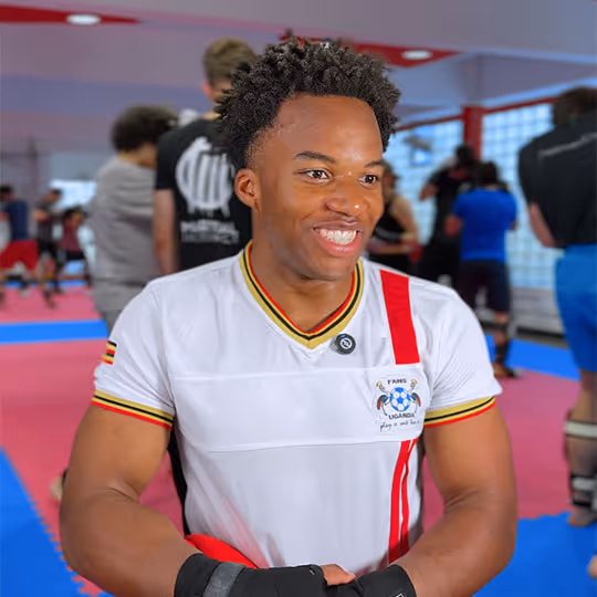 Junger Mann mit weißen Sporthandschuhen und weißem T-Shirt mit Fußballlogo, der in einer Kampfsporthalle lächelt.