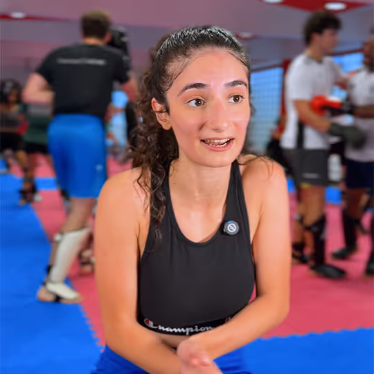Junge Frau in Sportkleidung in einem Boxstudio mit anderen Sportlern im Hintergrund.