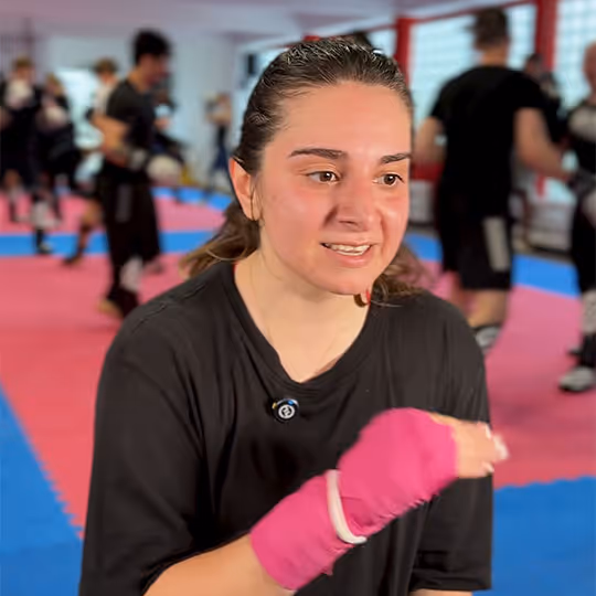Frau beim Boxtraining mit pinken Bandagen in einem Fitnessstudio mit anderen sportlichen Teilnehmern im Hintergrund.