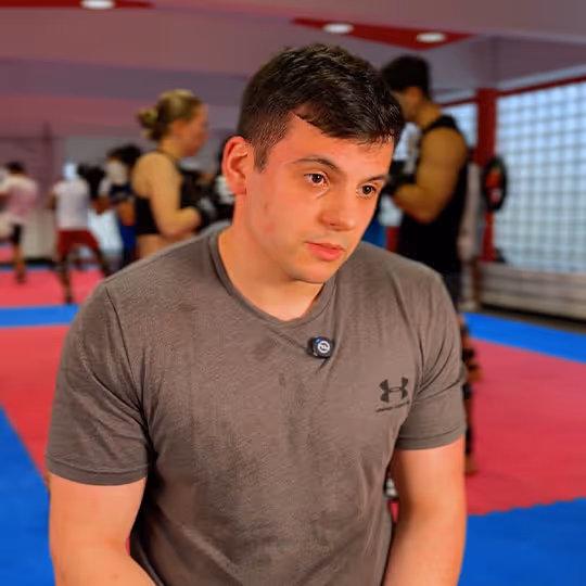 Junger Mann mit feuchtem grauem T-Shirt in einem Fitnessstudio mit anderen trainierenden Personen im Hintergrund.