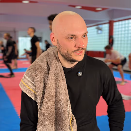 Mann mit Glatze und Bart trägt ein schwarzes Oberteil und hat ein Handtuch über die Schulter in einem Fitnessstudio.