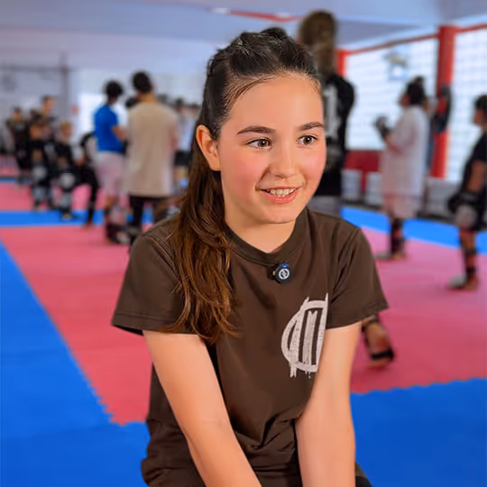 Mädchen mit braunem T-Shirt sitzt auf blauen und roten Matten in einem Kampfsportstudio.
