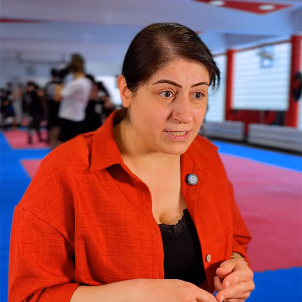 Frau mit rotem Hemd und schwarzem Unterhemd in einem Raum mit blauen und roten Matten, die spricht.