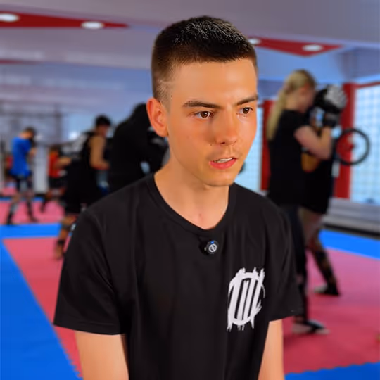 Junger Mann in schwarzem T-Shirt in einem Kampfsportstudio mit mehreren trainierenden Personen im Hintergrund.